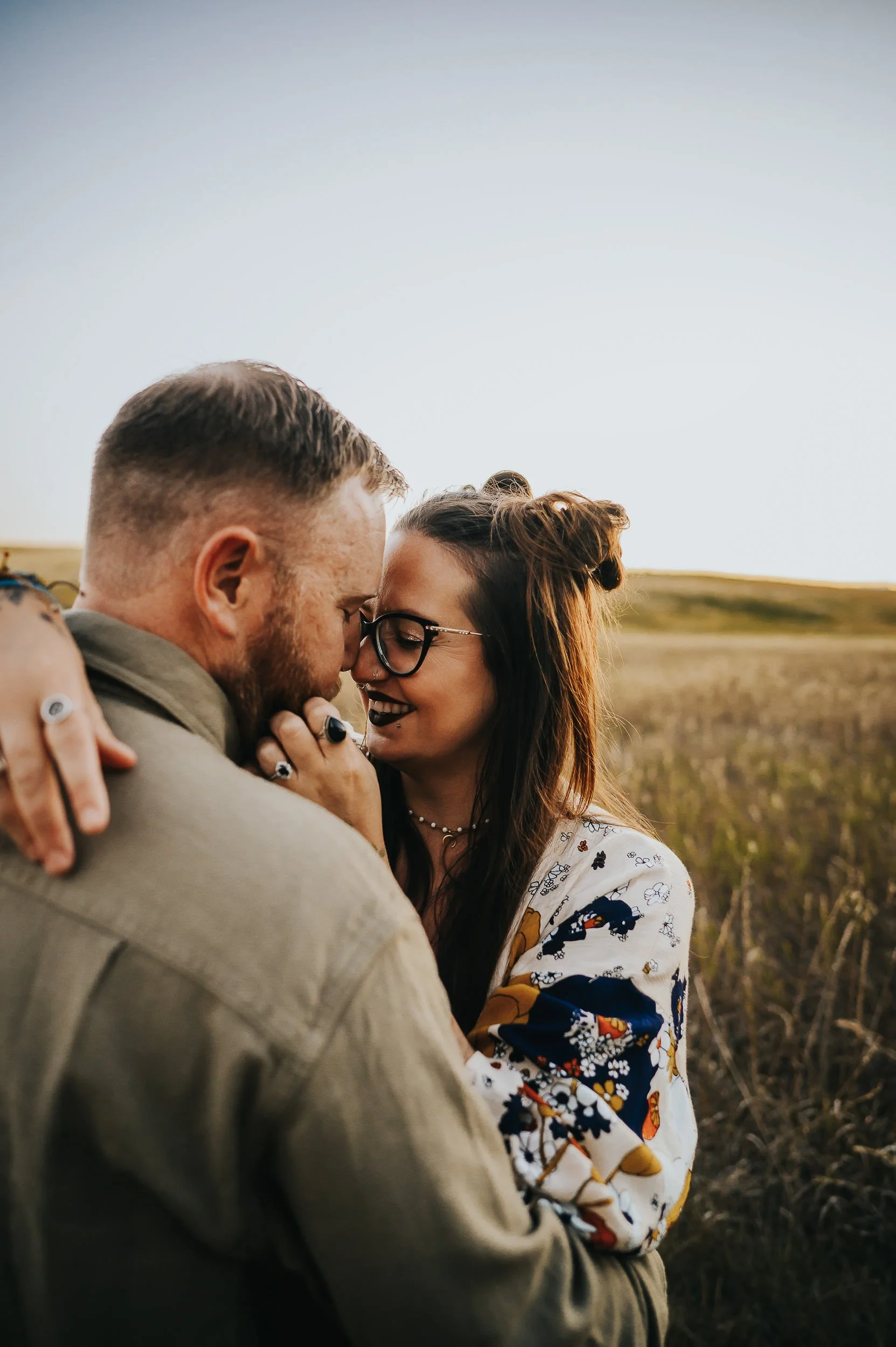 Close-up of couple, she wears round glasses and colorful floral top, he wears olive jacket and whispers to her as she smiles.
