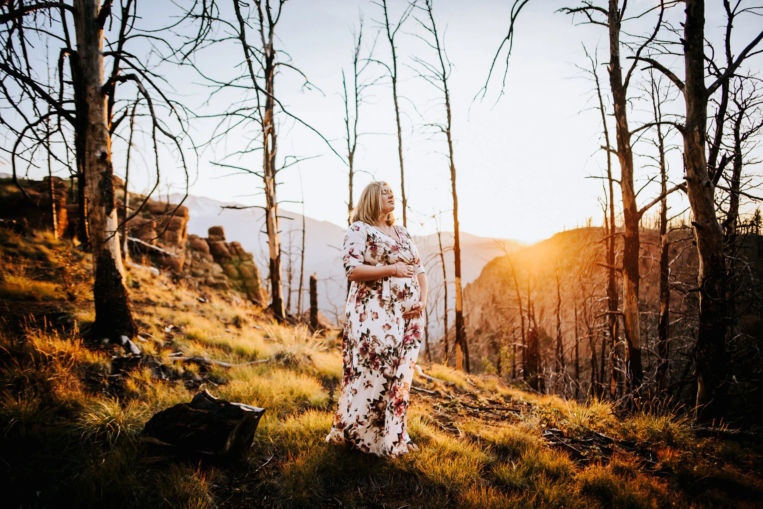 Pregnant woman in a white lace dress standing on a golden sunlit hillside surrounded by bare trees.