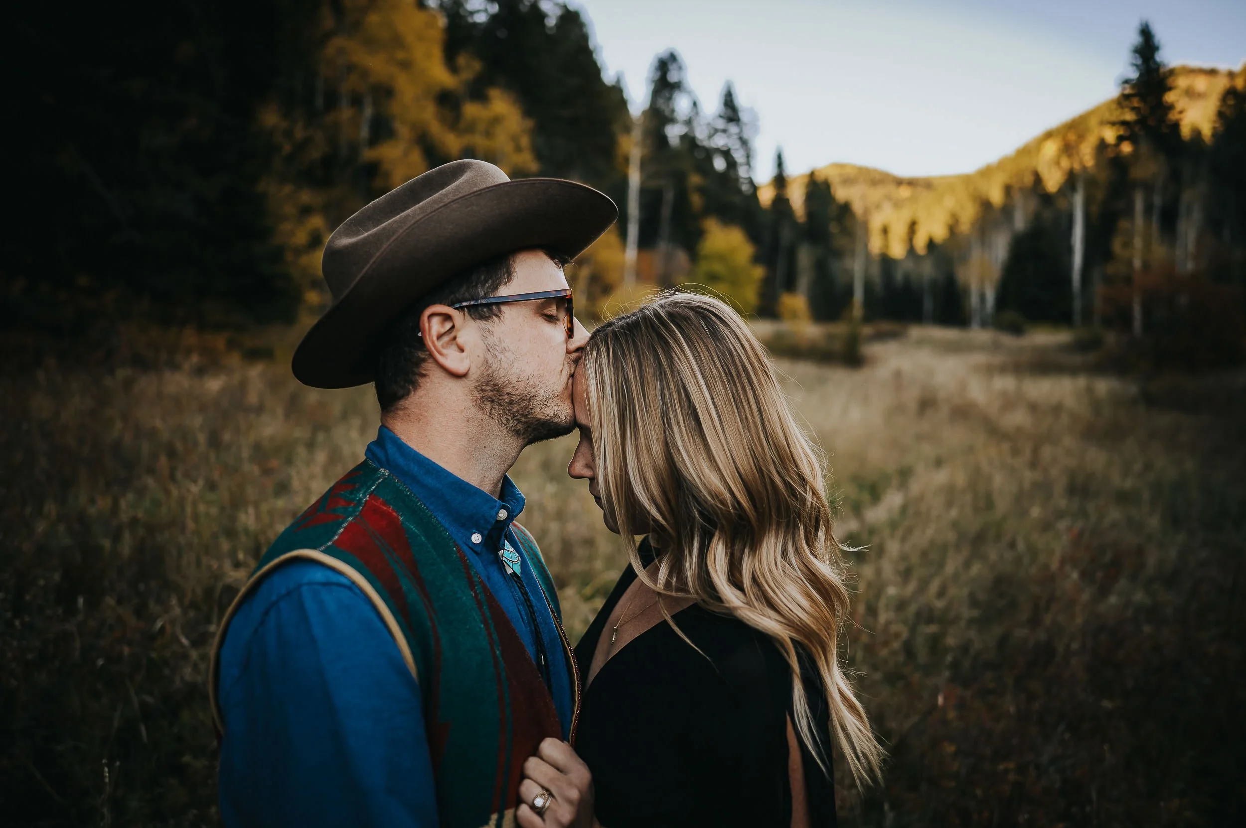 Couple in fall mountain setting with golden aspens, he wears hat and olive jacket, she has blonde hair in lighter earth tones.