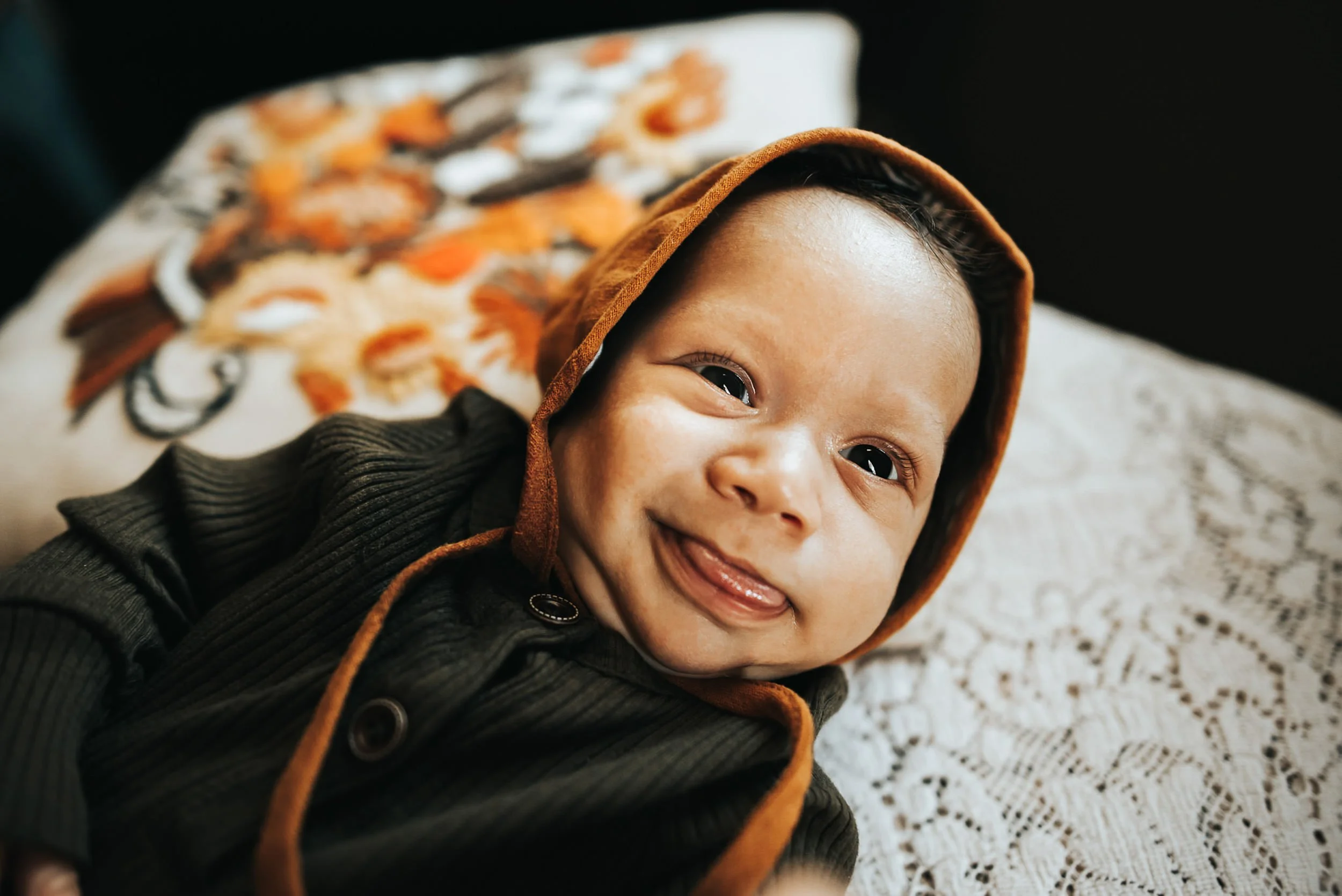 A newborn in a rust-colored bonnet looks up with a tiny open-mouth smile, closeup.