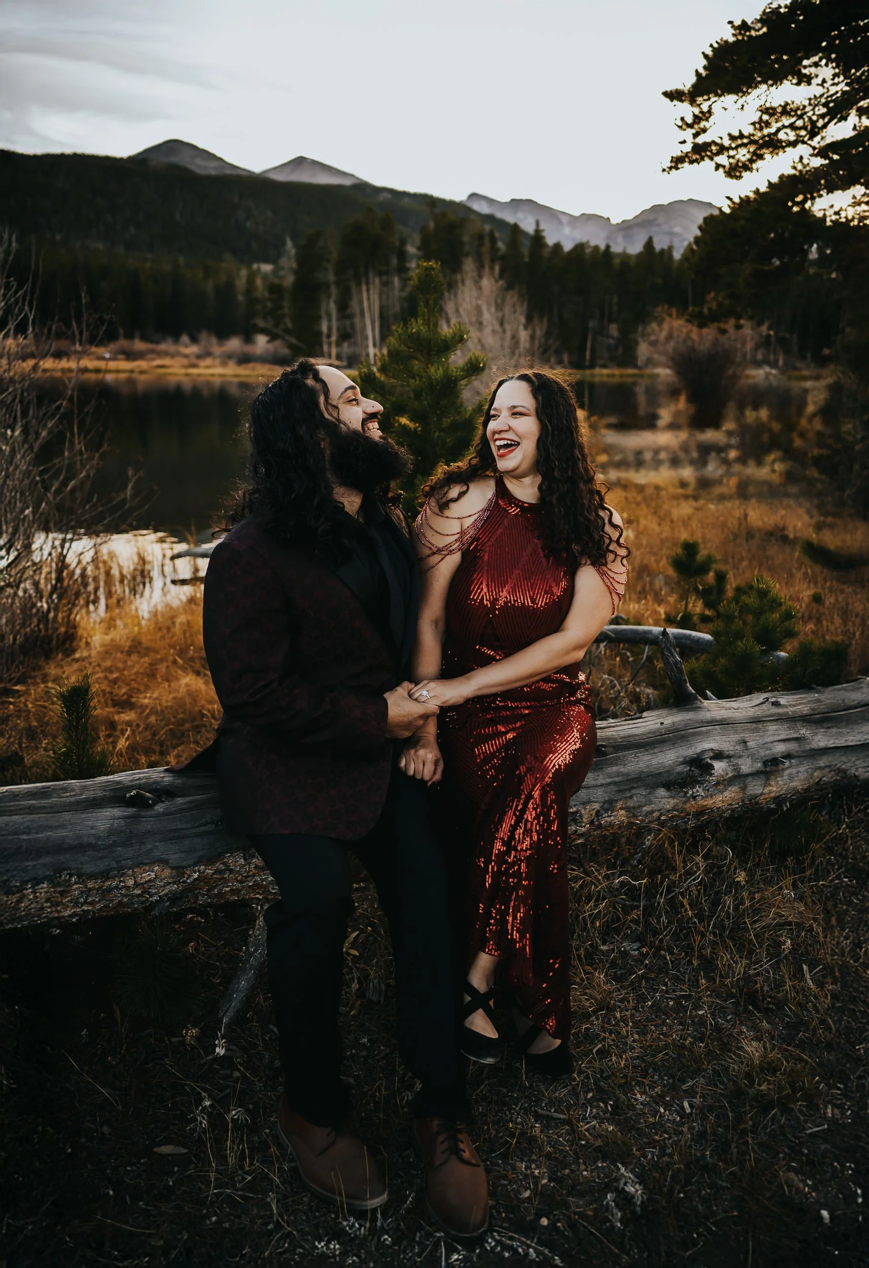 A couple in formal wear laughs and dances together on a log by a Colorado lake at dusk.