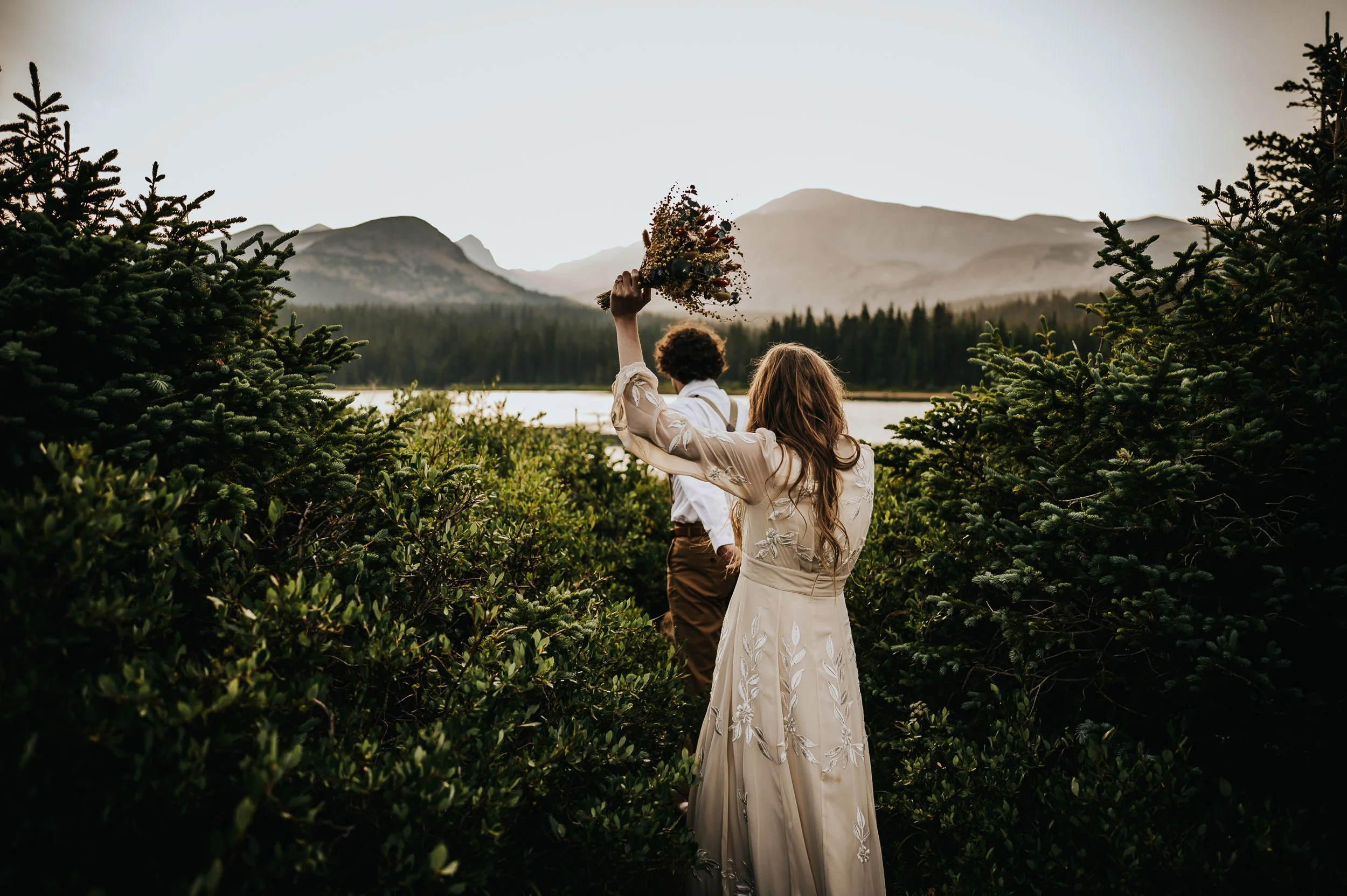 A bride raises her bouquet overhead by a Colorado mountain lake, her back to the camera, lace dress trailing.