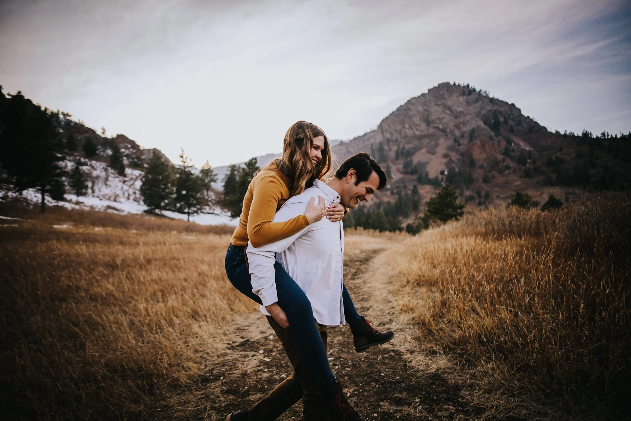 A couple dances together in a golden Colorado hillside field, she in a mustard top, both laughing.