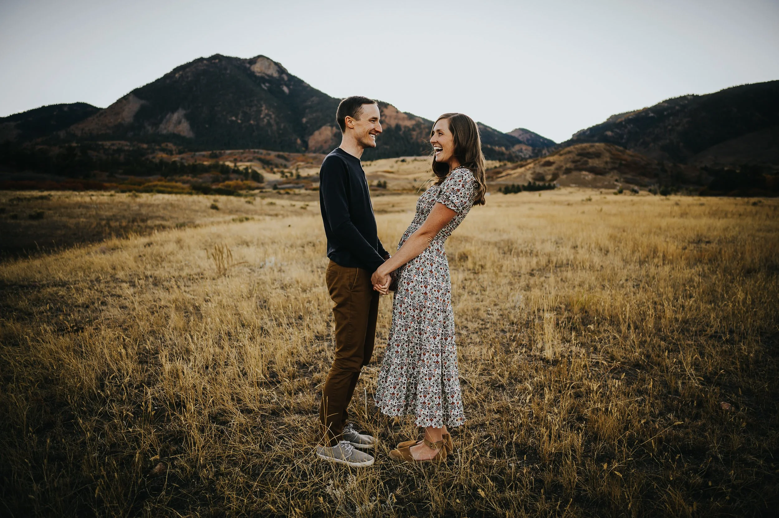 A couple stands facing each other in a wide Colorado mountain field, she in a floral dress, relaxed and connected.