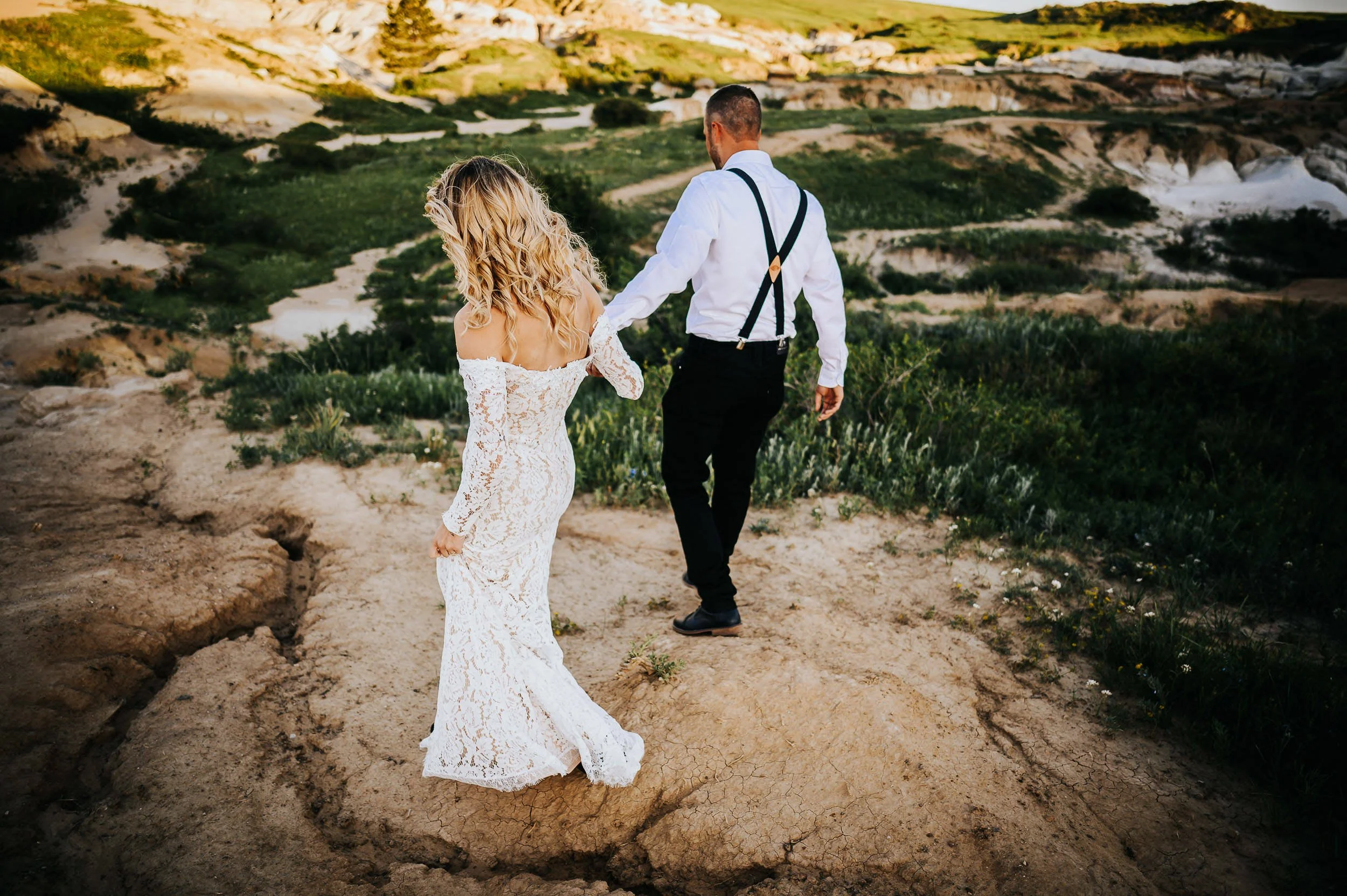 A bride and groom walk together across a colorful mineral landscape, she in a lace gown with a long train.