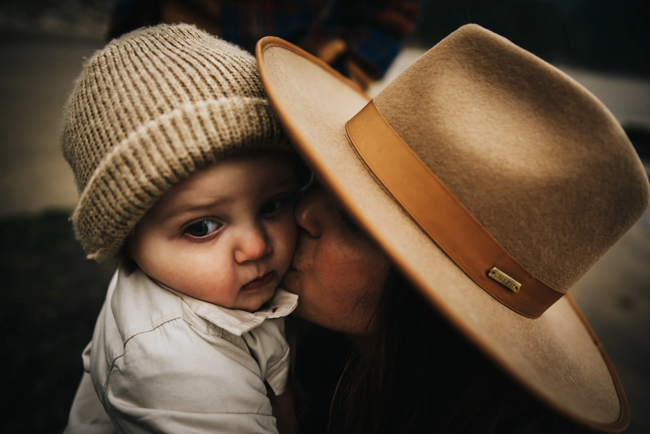 A baby is kissed on the cheek by his mother who is holding his close and wearing a wide brimmed hat with a goldenrod hat band.