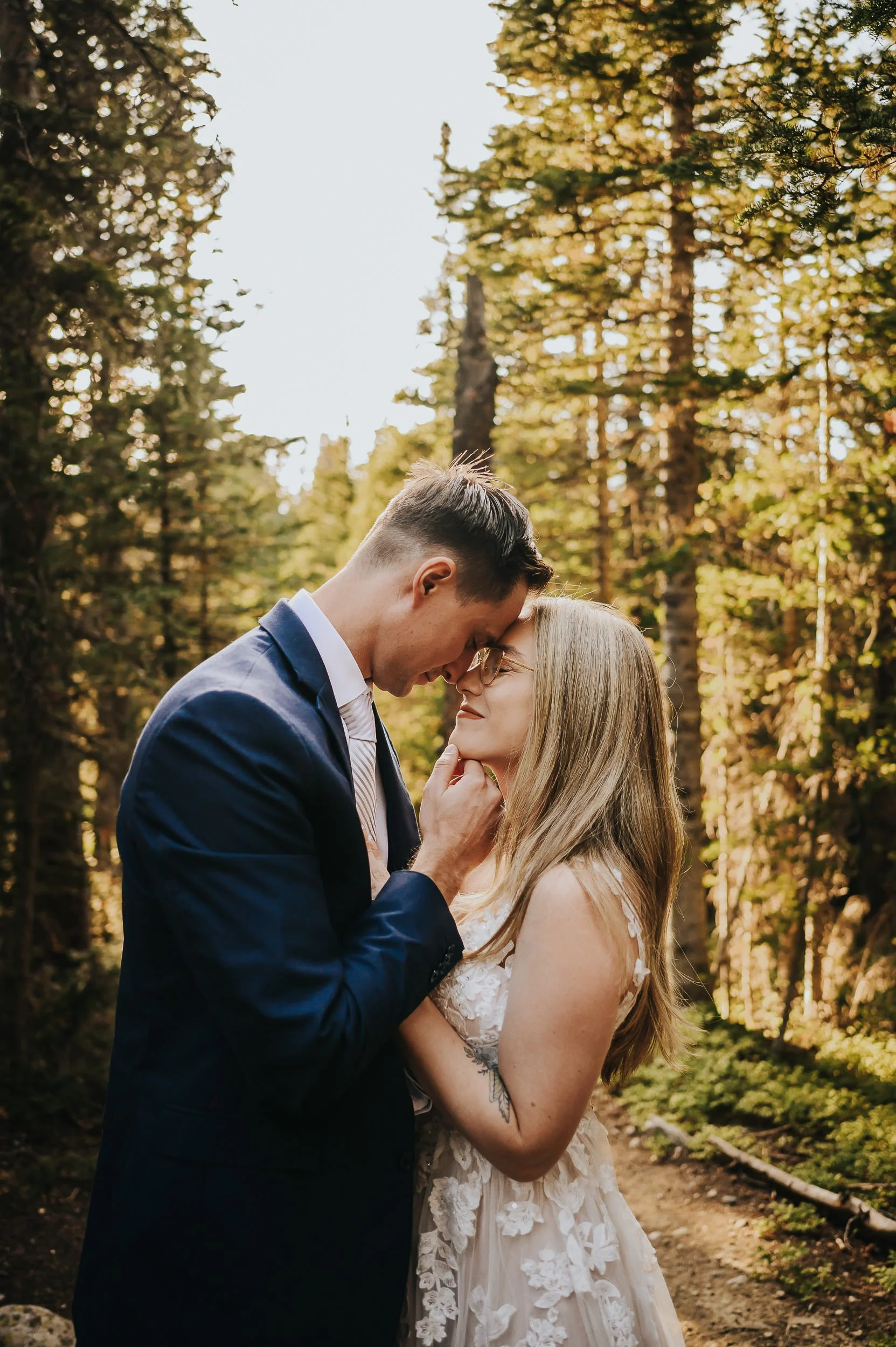 A couple shares a close forehead-to-forehead moment in a sunlit Colorado pine forest, golden light all around them.