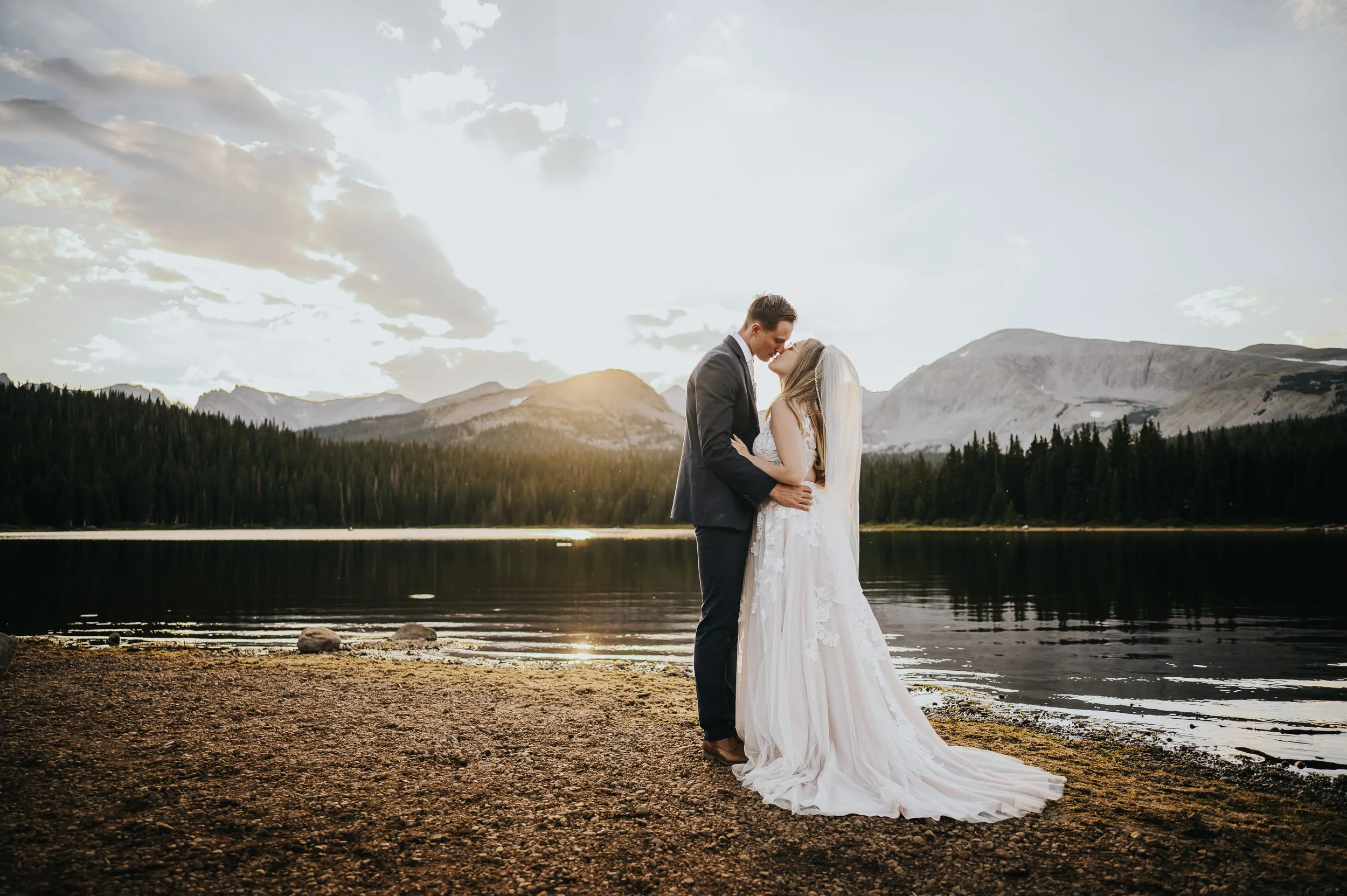 A bride and groom stand together at the edge of a mountain lake at sunset, soft golden light behind them.
