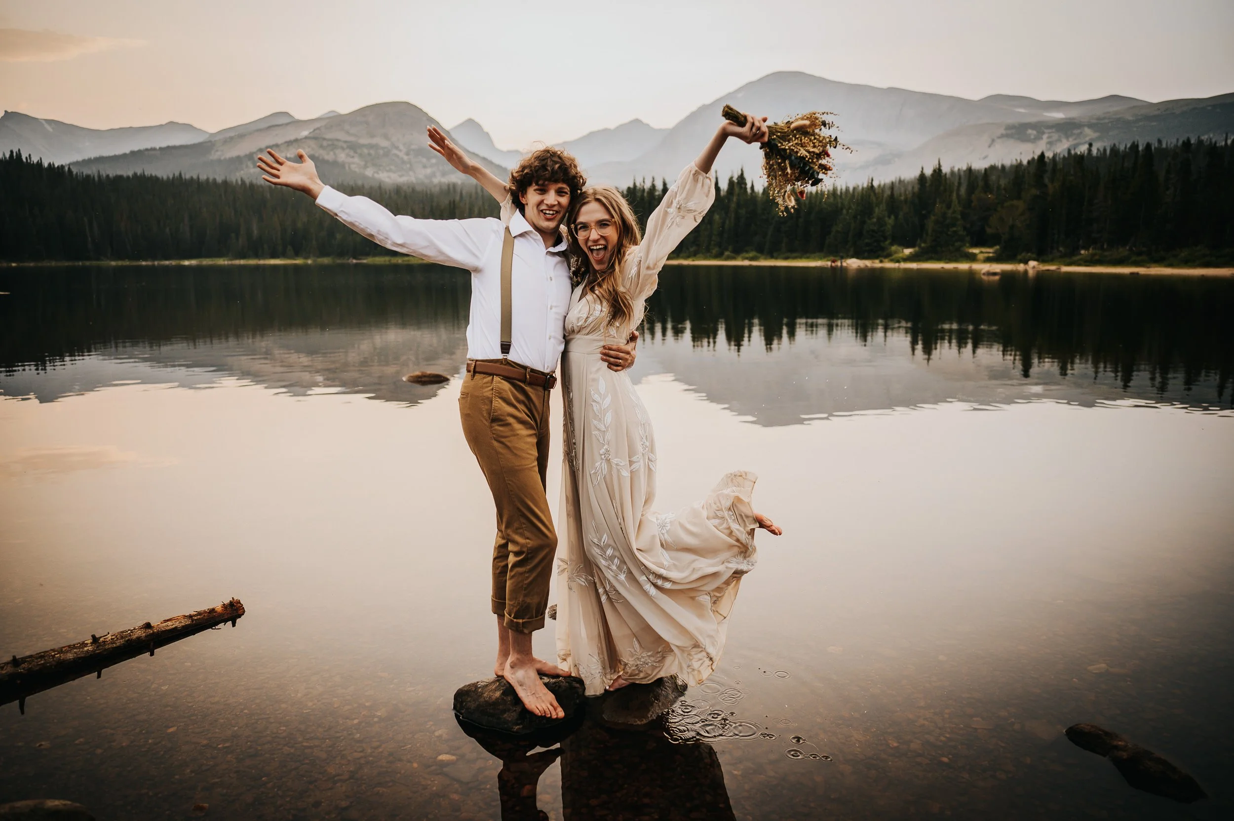A bride throws her bouquet overhead by a mountain lake, groom beside her, celebrating their elopement at Brainard Lake near Ward, Colorado.