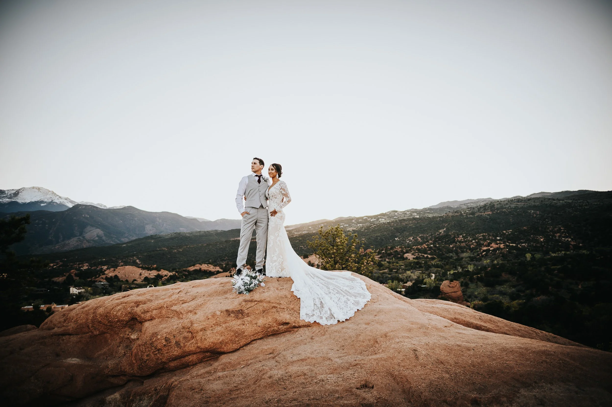 A bride and groom stand on a boulder together in a wide Colorado mountain landscape, full wedding attire at Garden of the Gods after their wedding ceremony at The Pinery on the Hill in Colorado Springs.