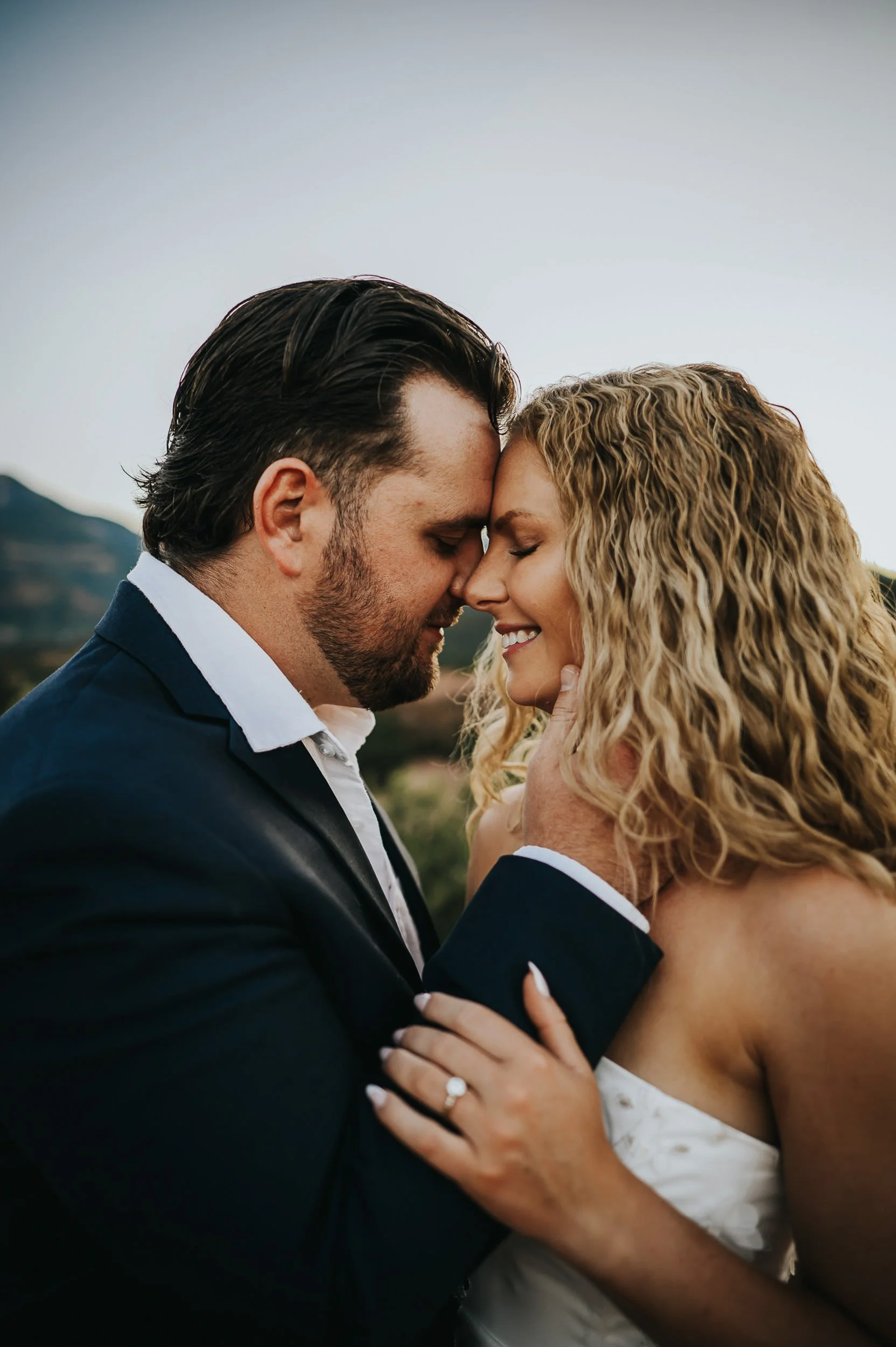 A bride and groom press foreheads together, both in wedding attire, soft emotional portrait.