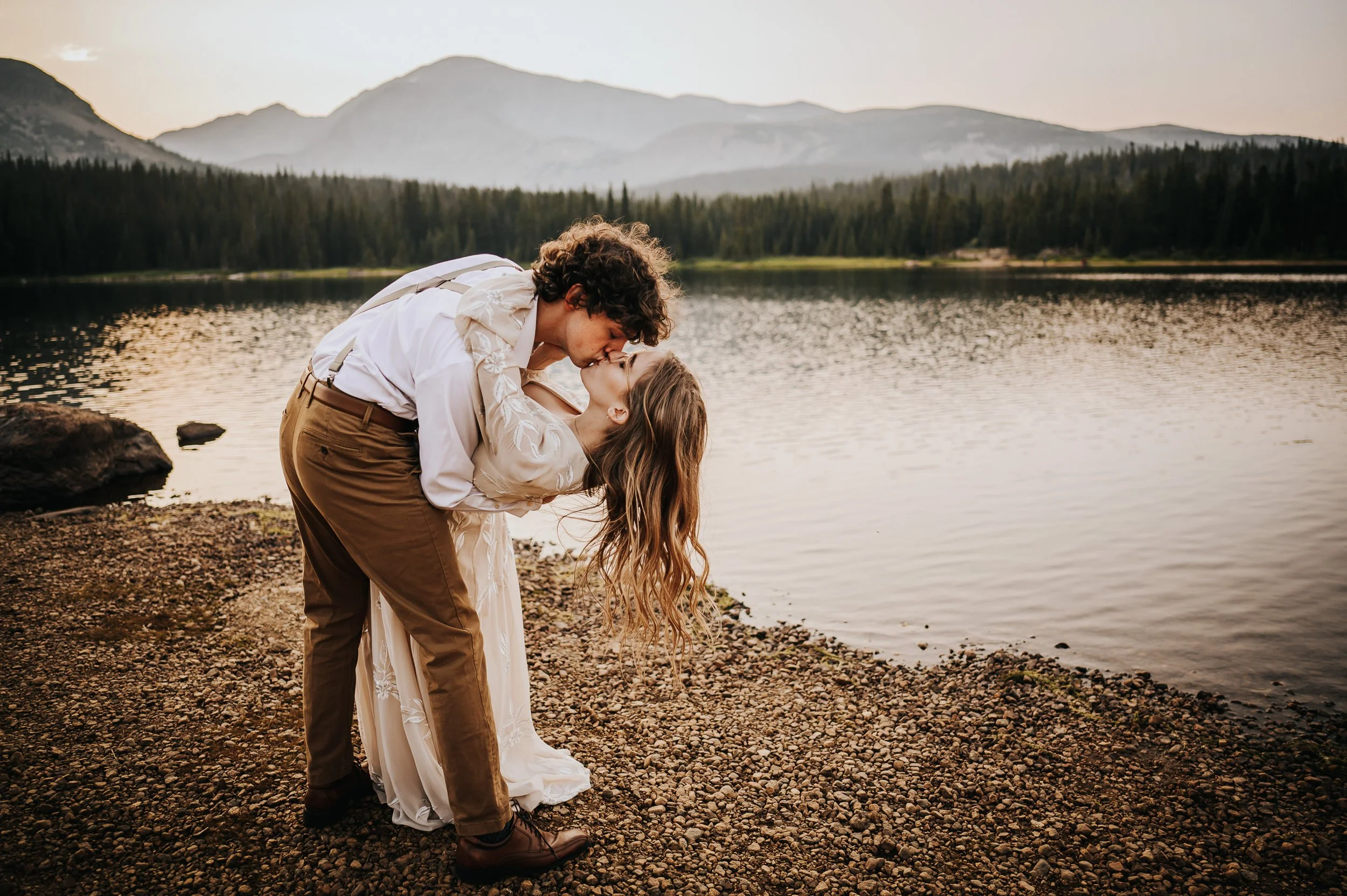 A groom dips his bride near the shore of a mountain lake, her curly hair falling toward the water.