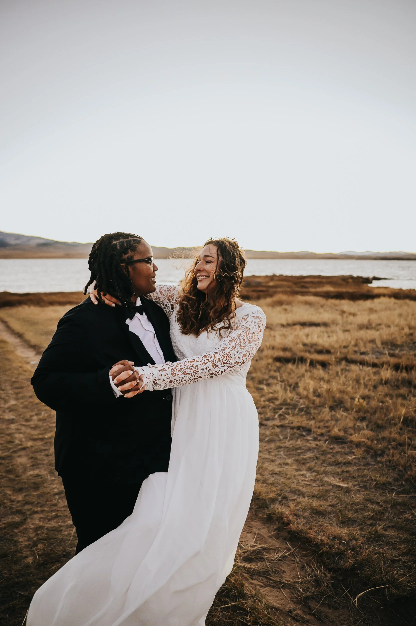 Two brides laughing and dancing together in a golden field with mountain views during their Front Range elopement.