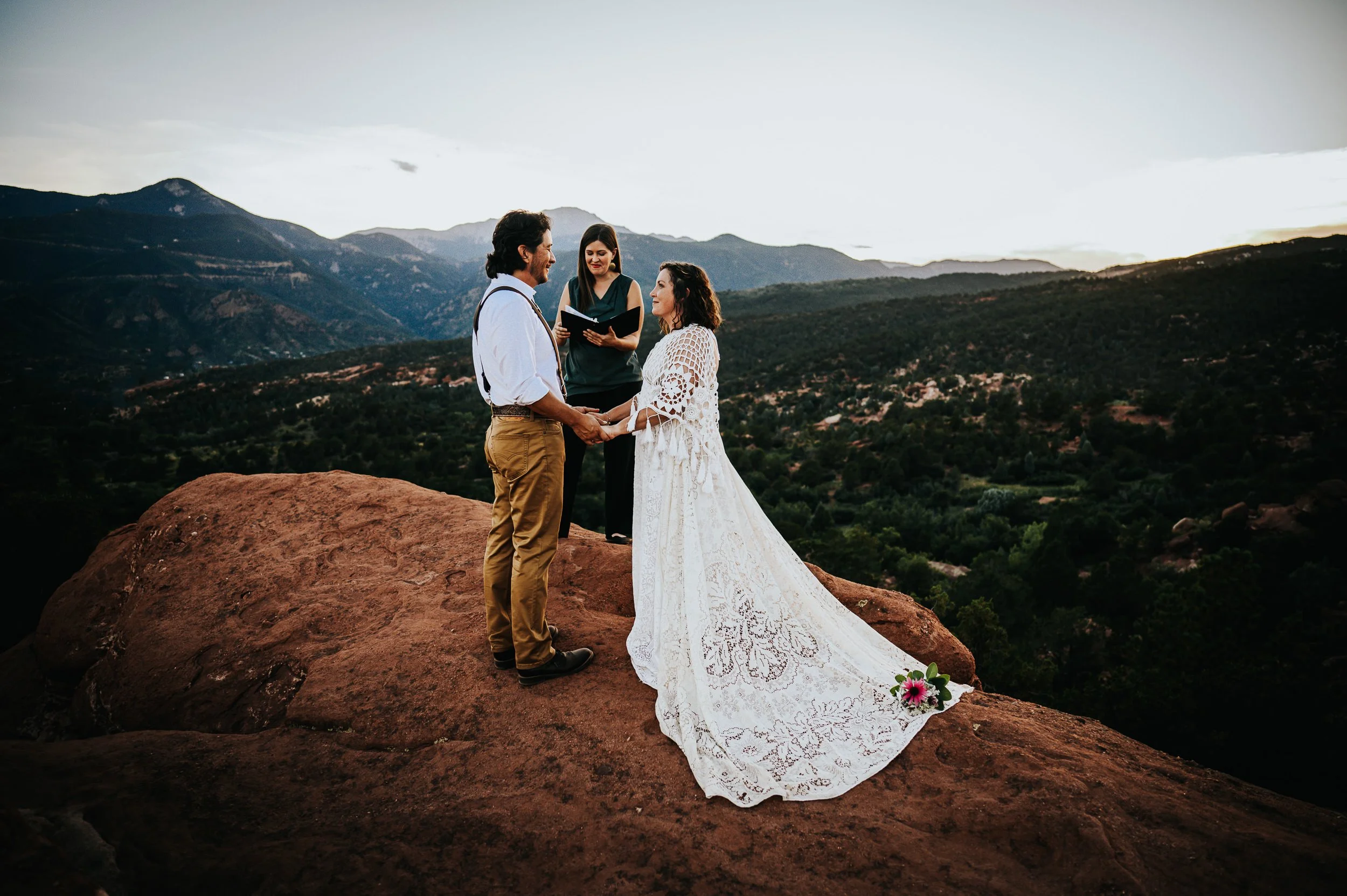 A bride and groom stand on a red rock overlook with their officiant, Colorado mountain panorama stretching behind them.