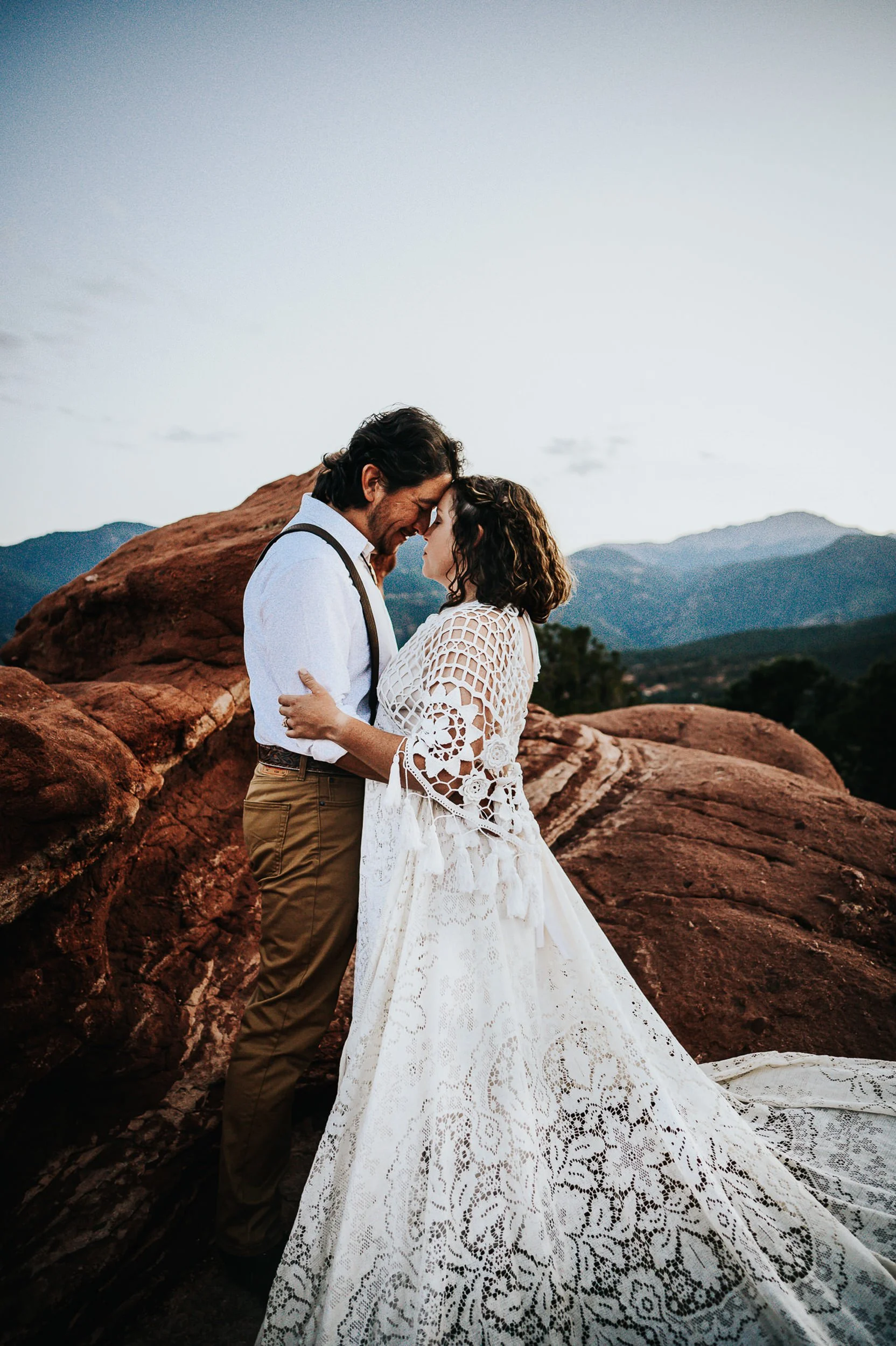 A couple shares a kiss on red rocks, she in a long lace gown, he in suspenders, Colorado mountains behind them.