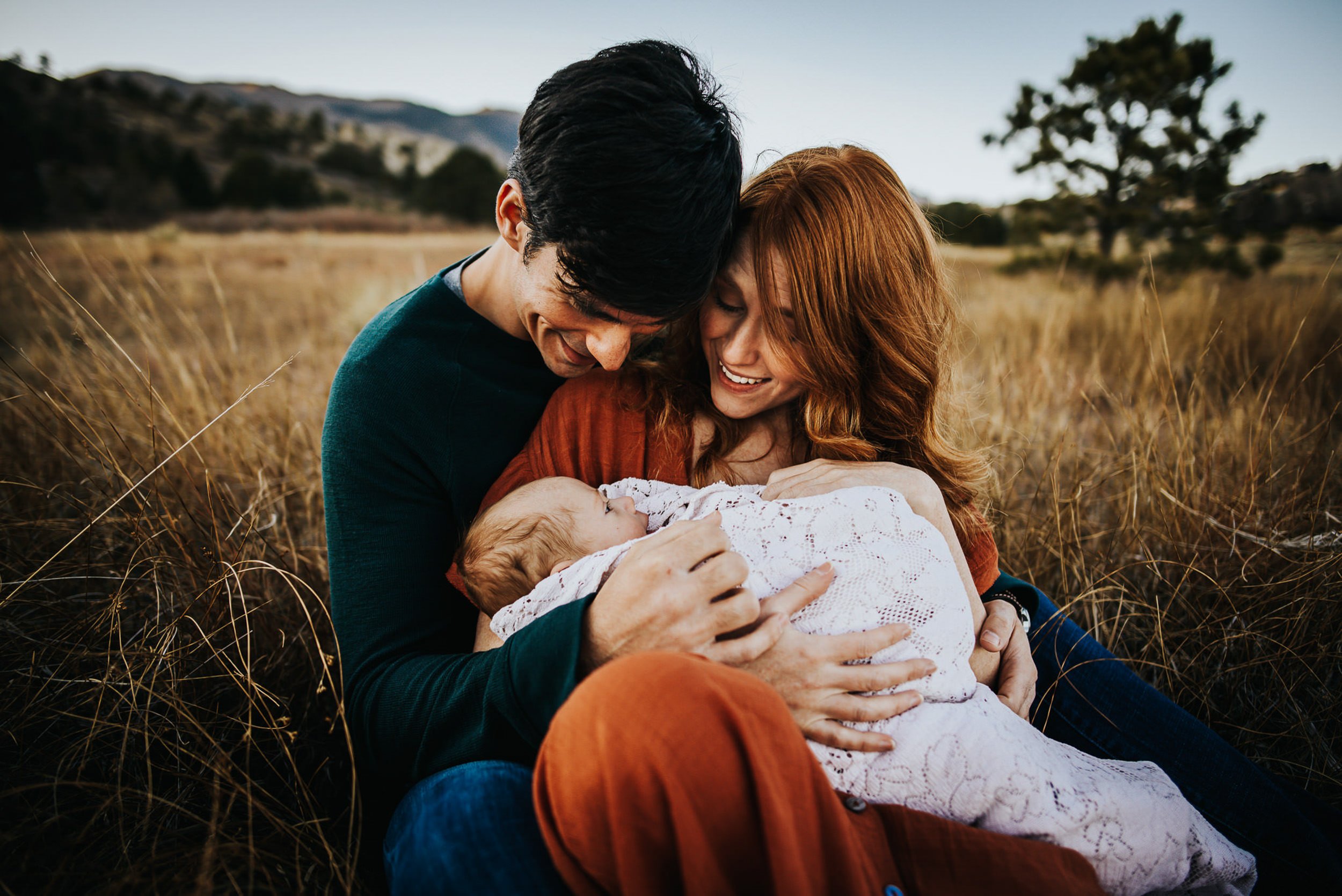 A couple sits together in a golden Colorado field cradling their newborn.