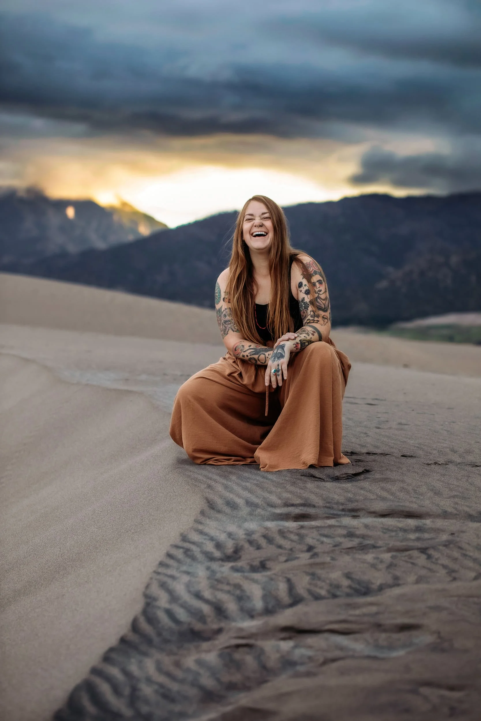 Sandy Patterson, Colorado photographer and educator kneels in the sand while laughing, tattoos showing at Great Sand Dunes National Park.