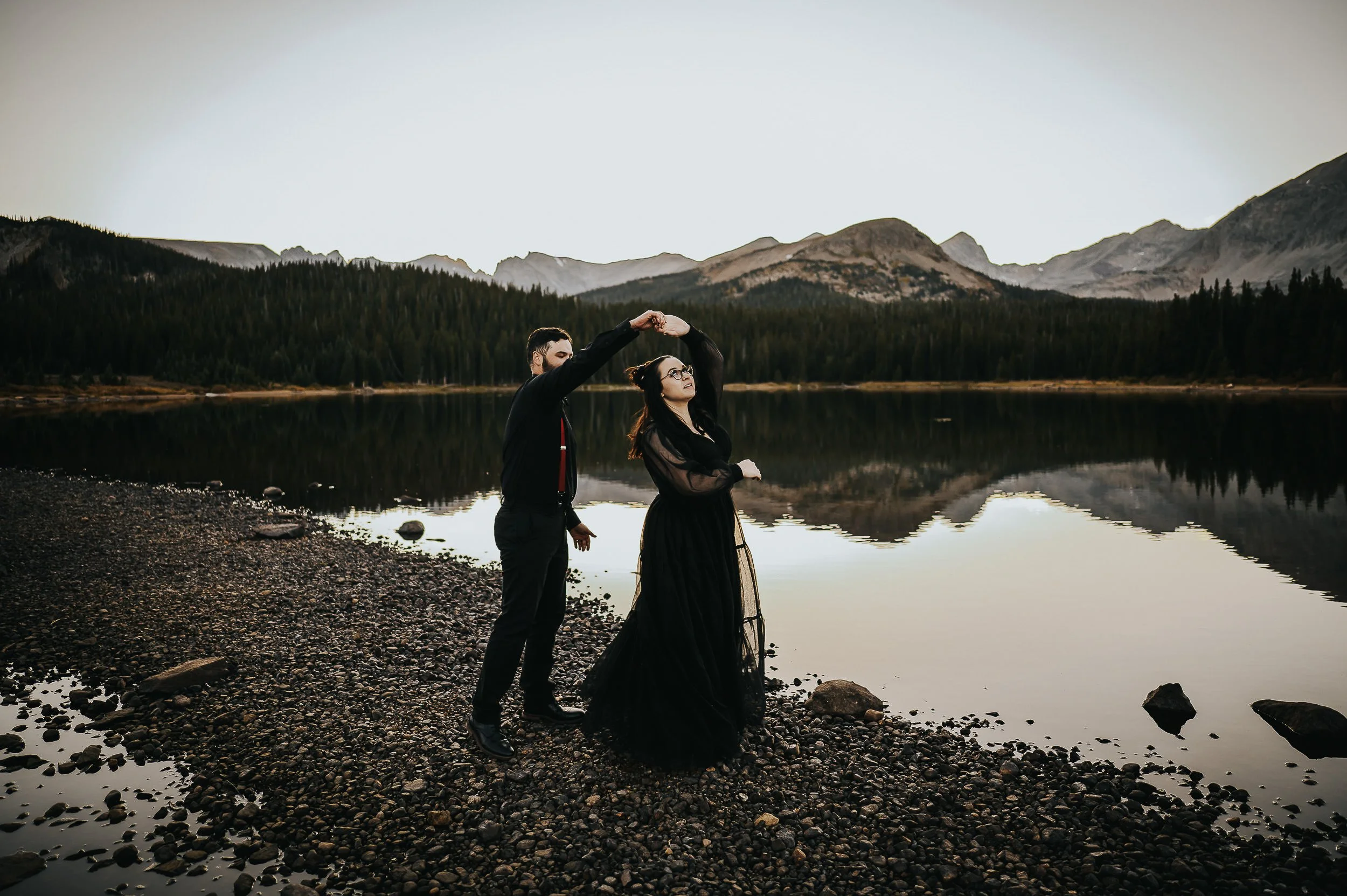 A woman in a black dress stands at the edge of a reflective alpine lake, her partner beside her, Colorado peaks behind them.