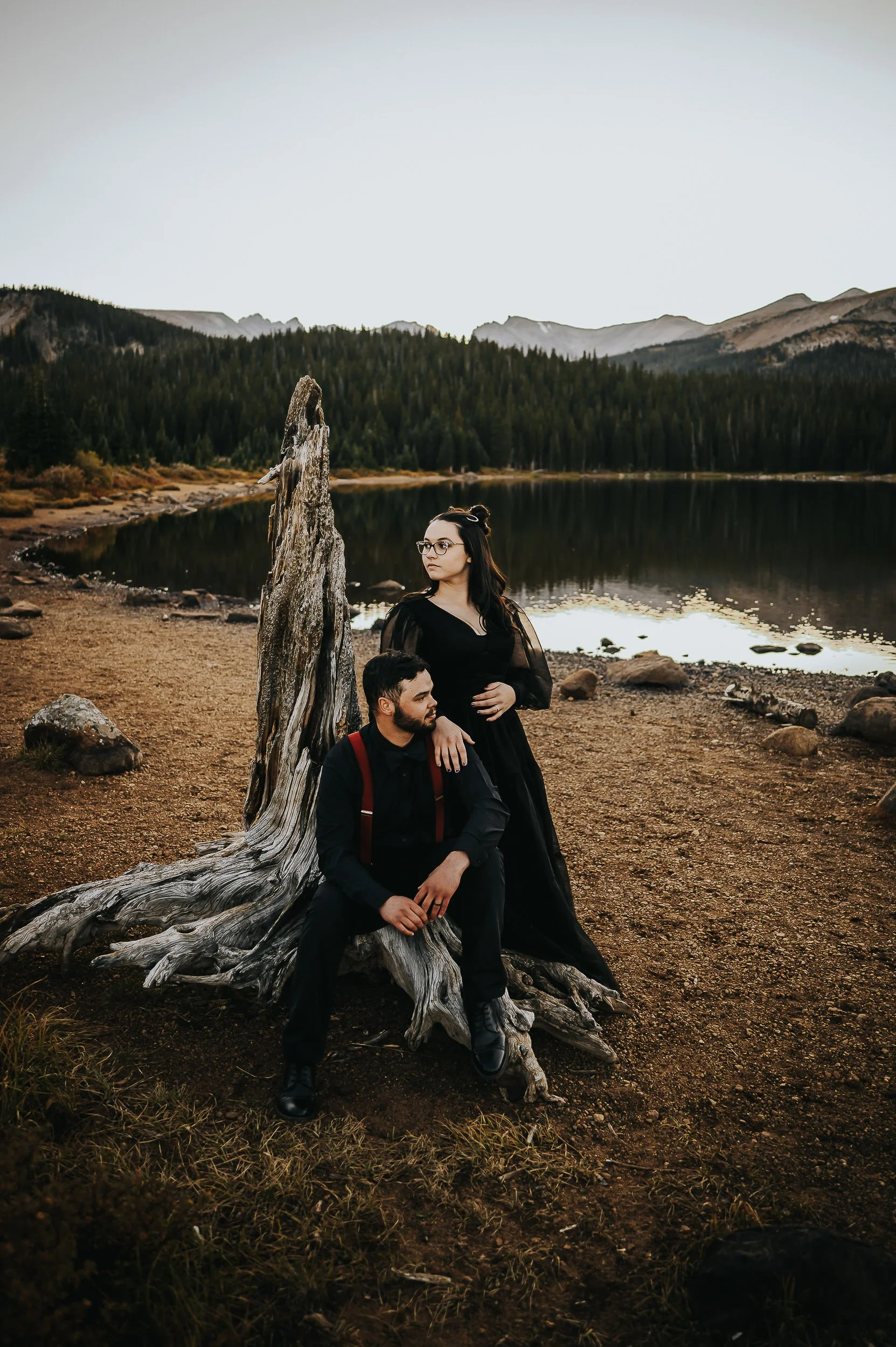 A woman in a black dress sits on a weathered log by a Colorado mountain lake, her partner standing beside her.