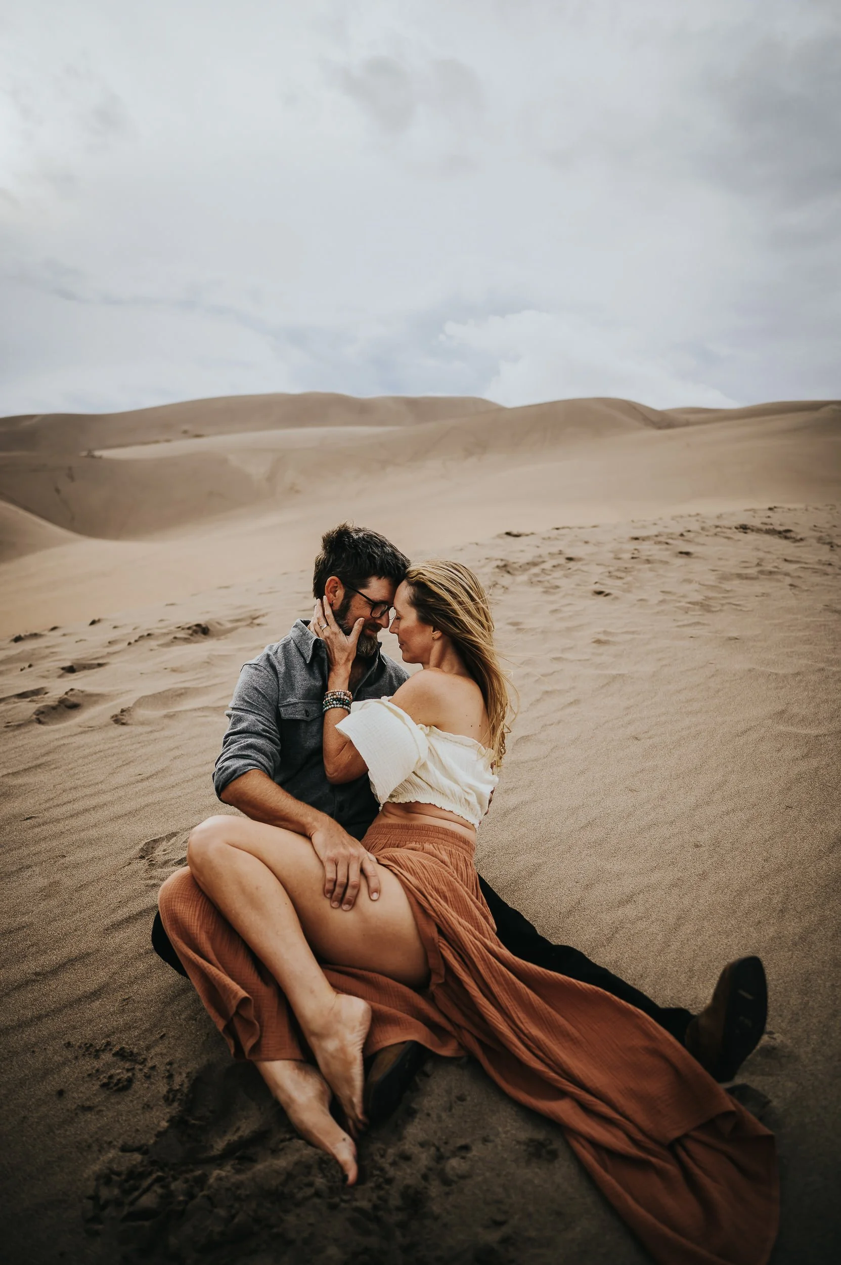 Couple in intimate embrace at Great Sand Dunes, she wears cream, sand dunes stretching behind them.