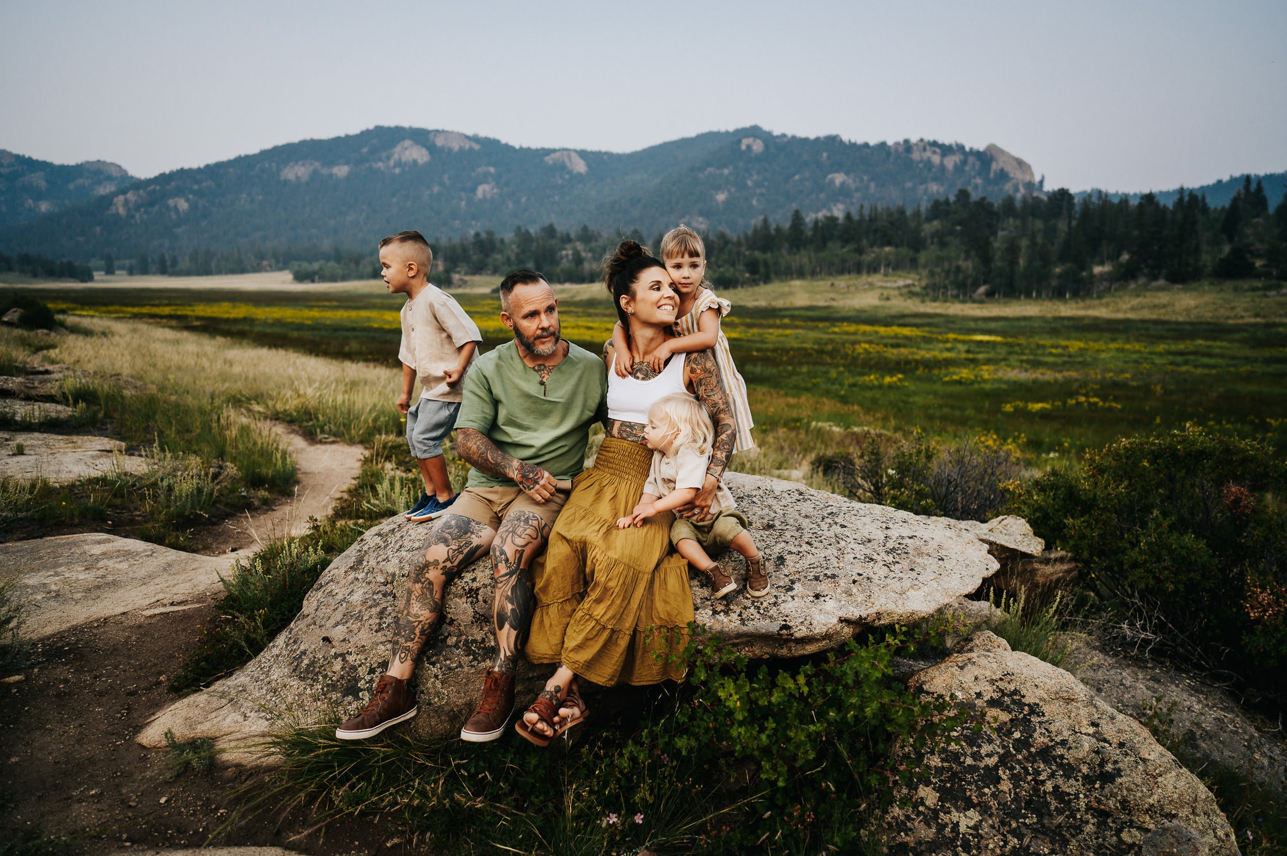 Family sitting on a large boulder near a high mountain meadow with wildflowers and distant peaks.
