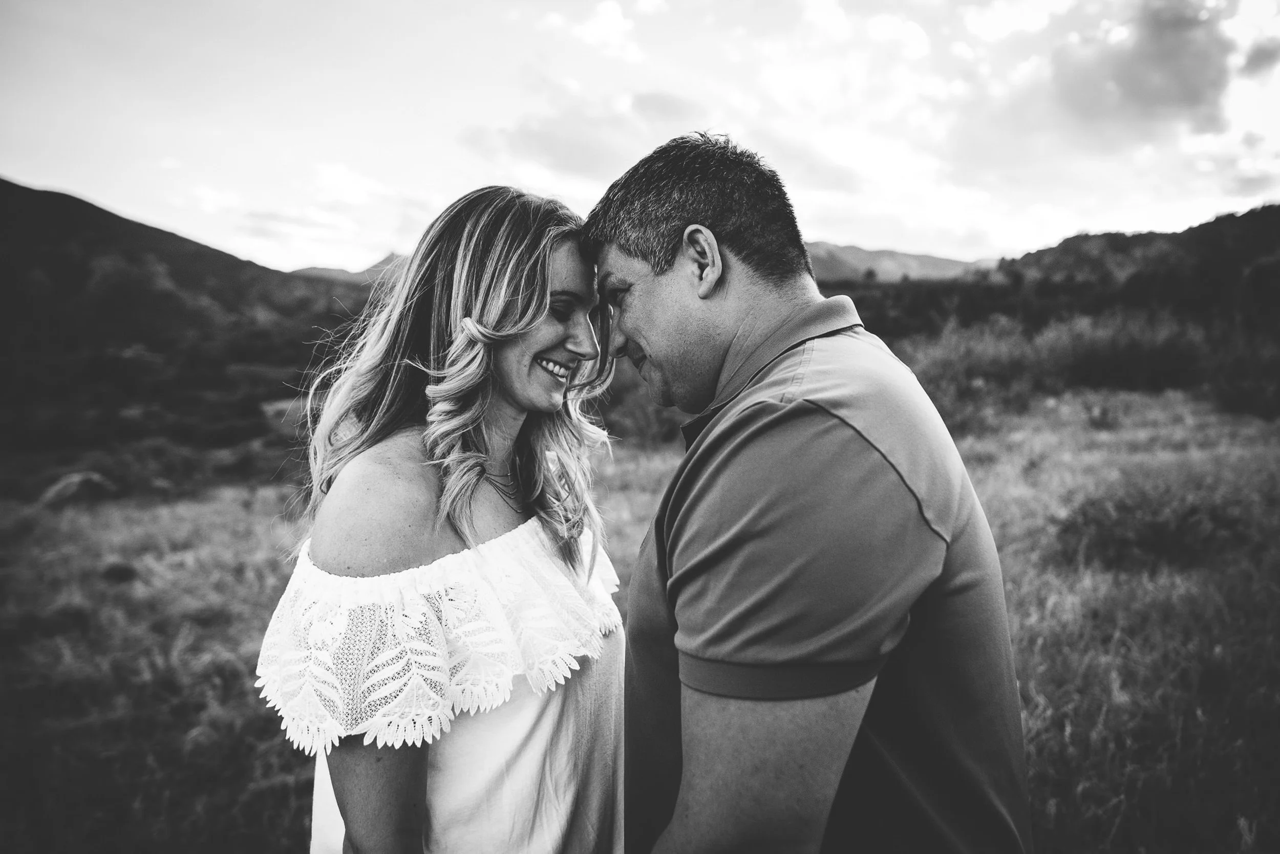 A couple leans forehead to forehead in a black and white portrait, her off-shoulder dress and his suspenders visible.