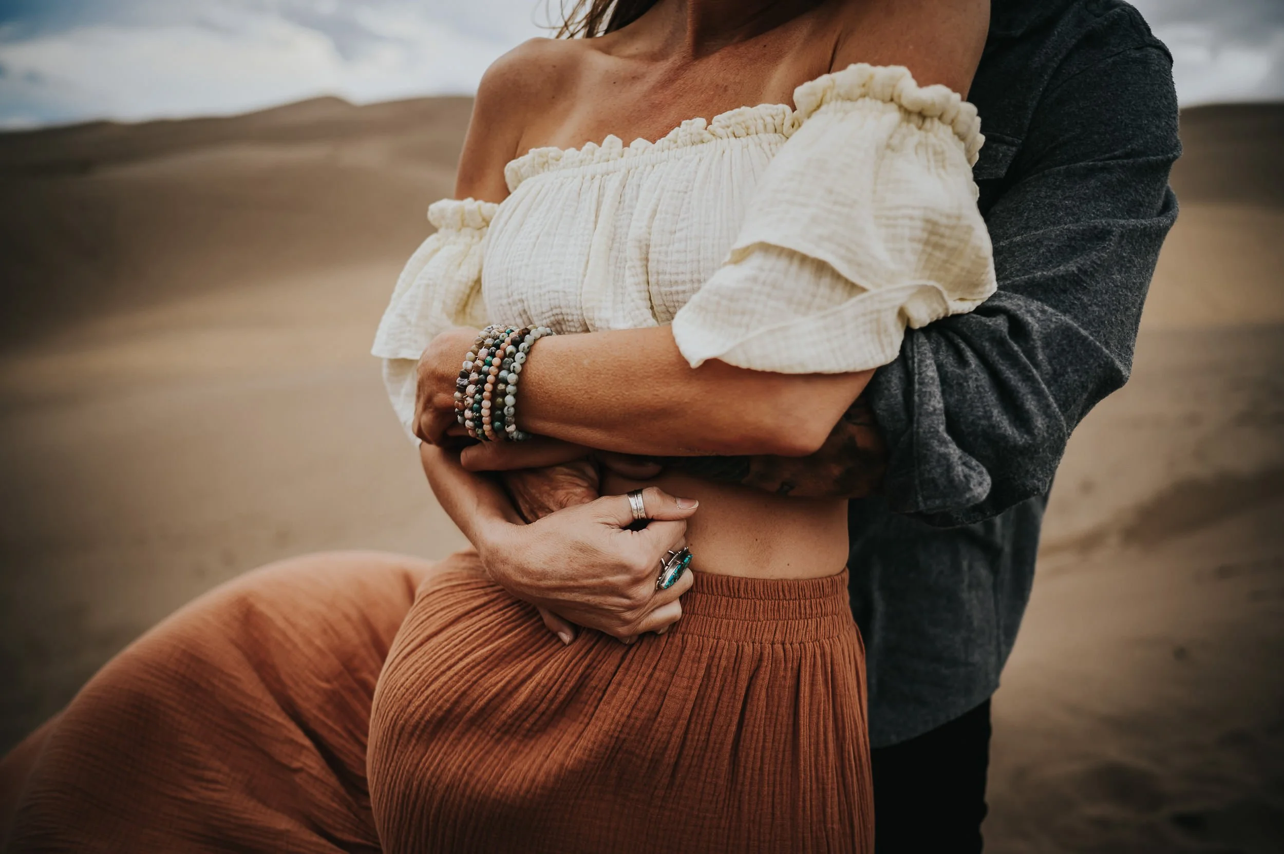 Close-up detail of couple embracing showing cream ruffled top, stacked beaded bracelets, chunky silver rings, and gray sweater sleeves  of her partner wrapped around her.