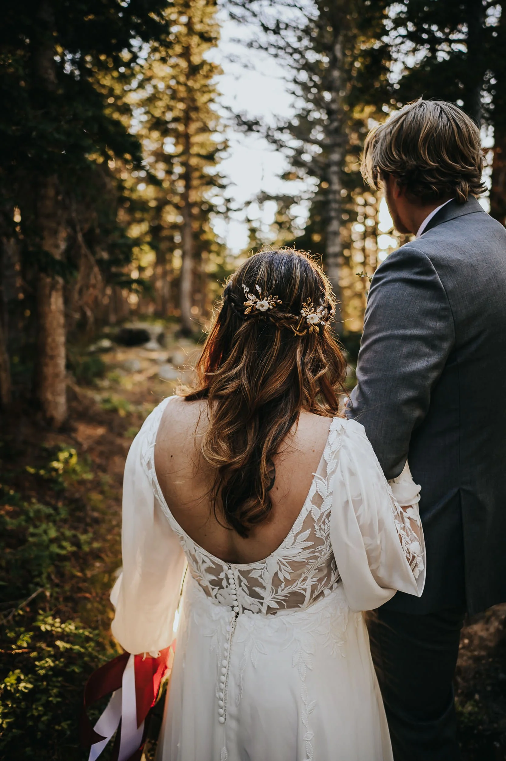 A bride with floral hair details walks ahead through a Colorado pine forest, her groom just behind her, her open lace back visible.