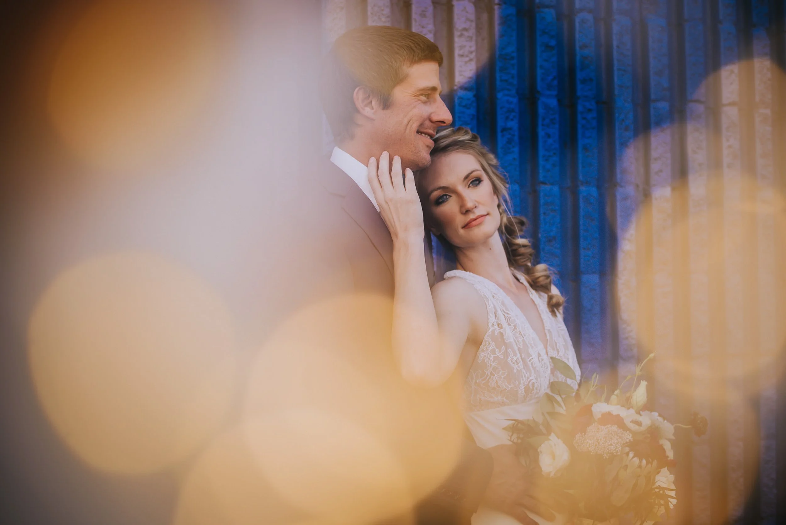 A bride leans back against her groom in dreamy golden backlight, blue barn door behind them.