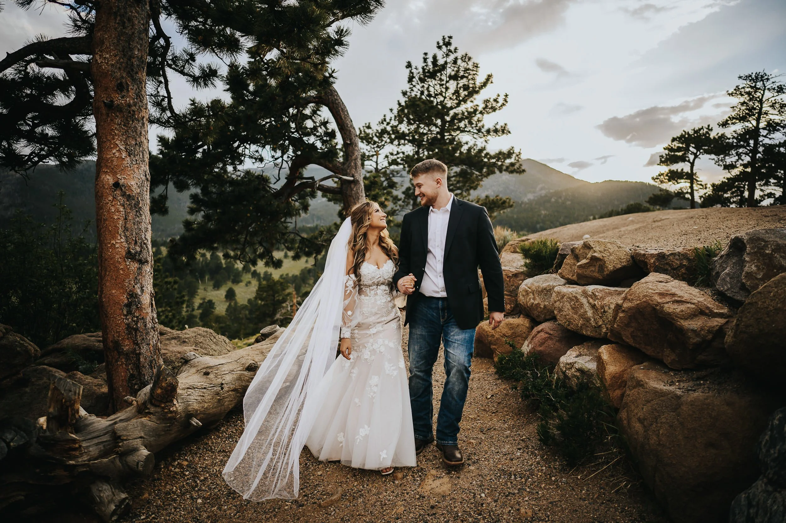 Bride in a lace gown with a long flowing veil walking a rocky trail with her groom, pine trees and mountains behind them.