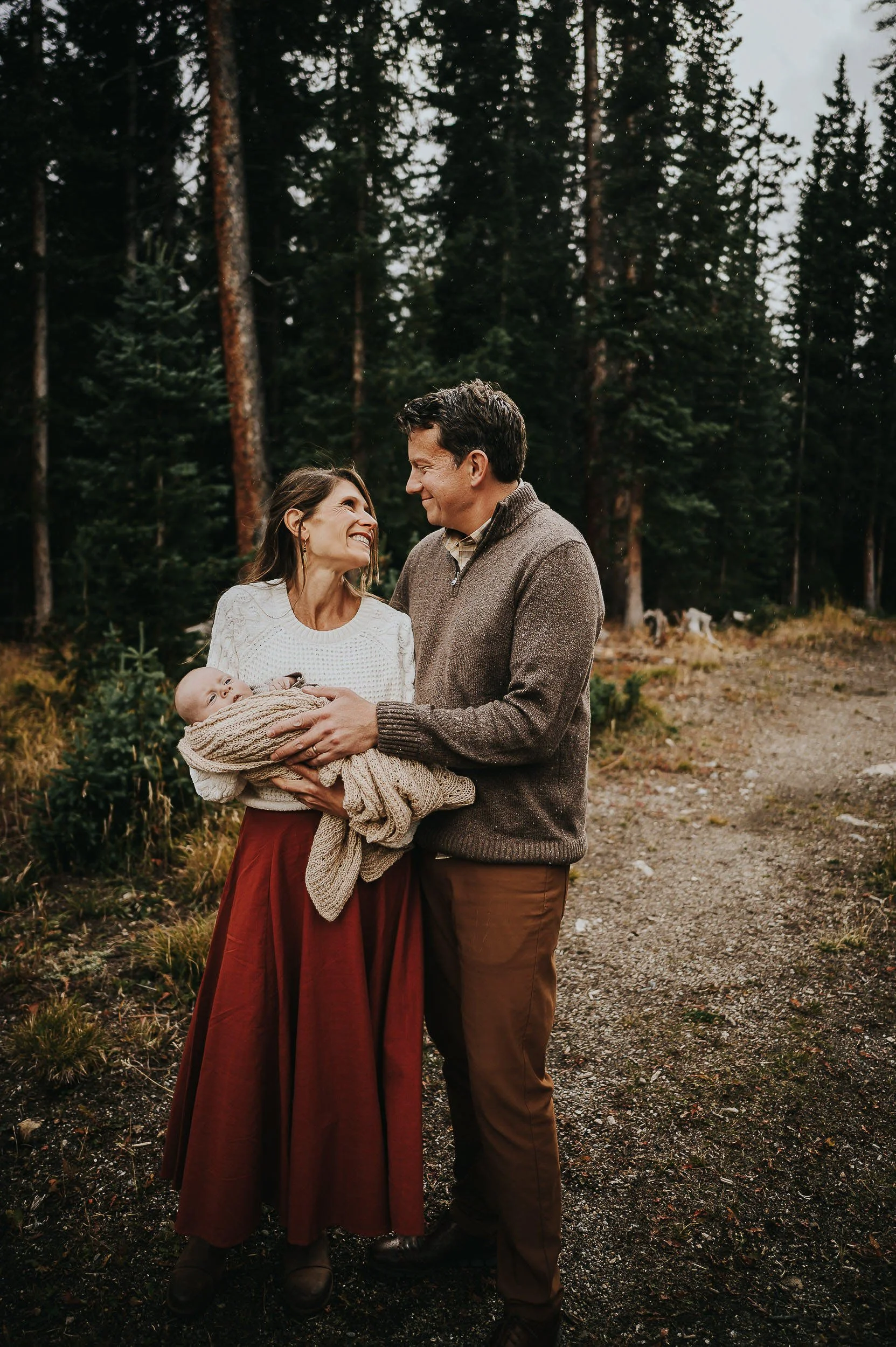 Couple standing together in forest clearing, mom in rust dress and dad in olive sweater holding their child, naturally coordinated.