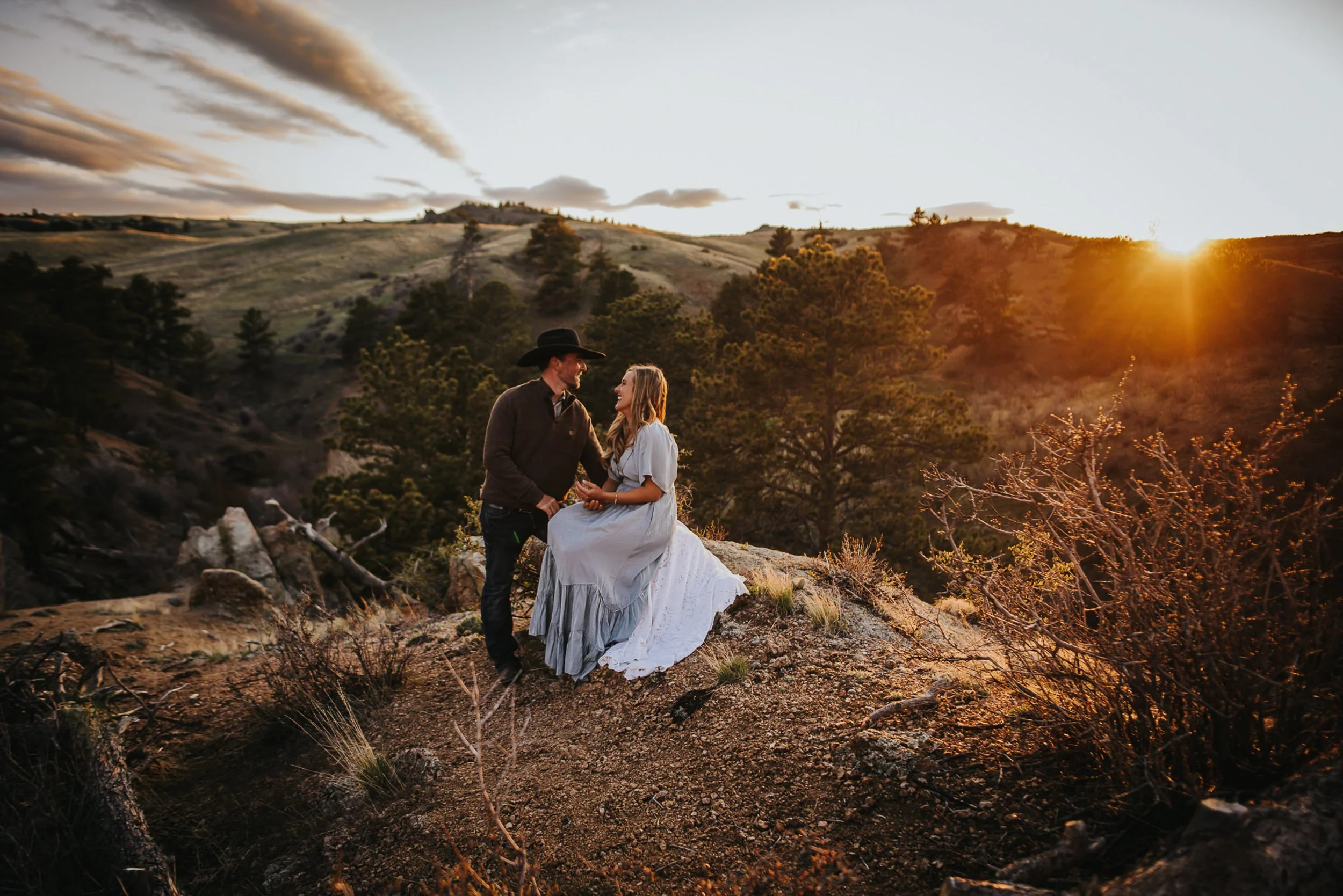 A couple sits close together on a rocky hillside at golden hour, Colorado mountains behind them.