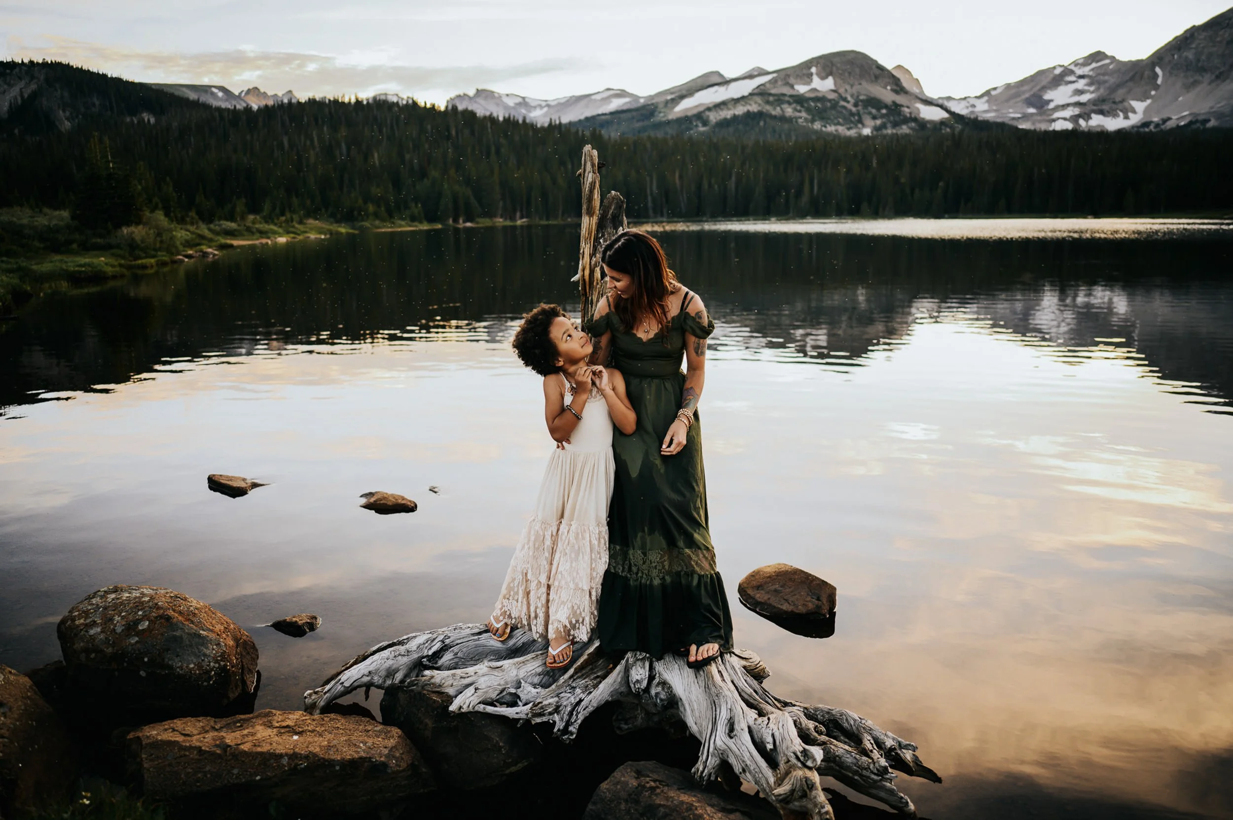 Woman in cream dress standing at an alpine lake with dramatic Colorado mountain backdrop and moody sky.