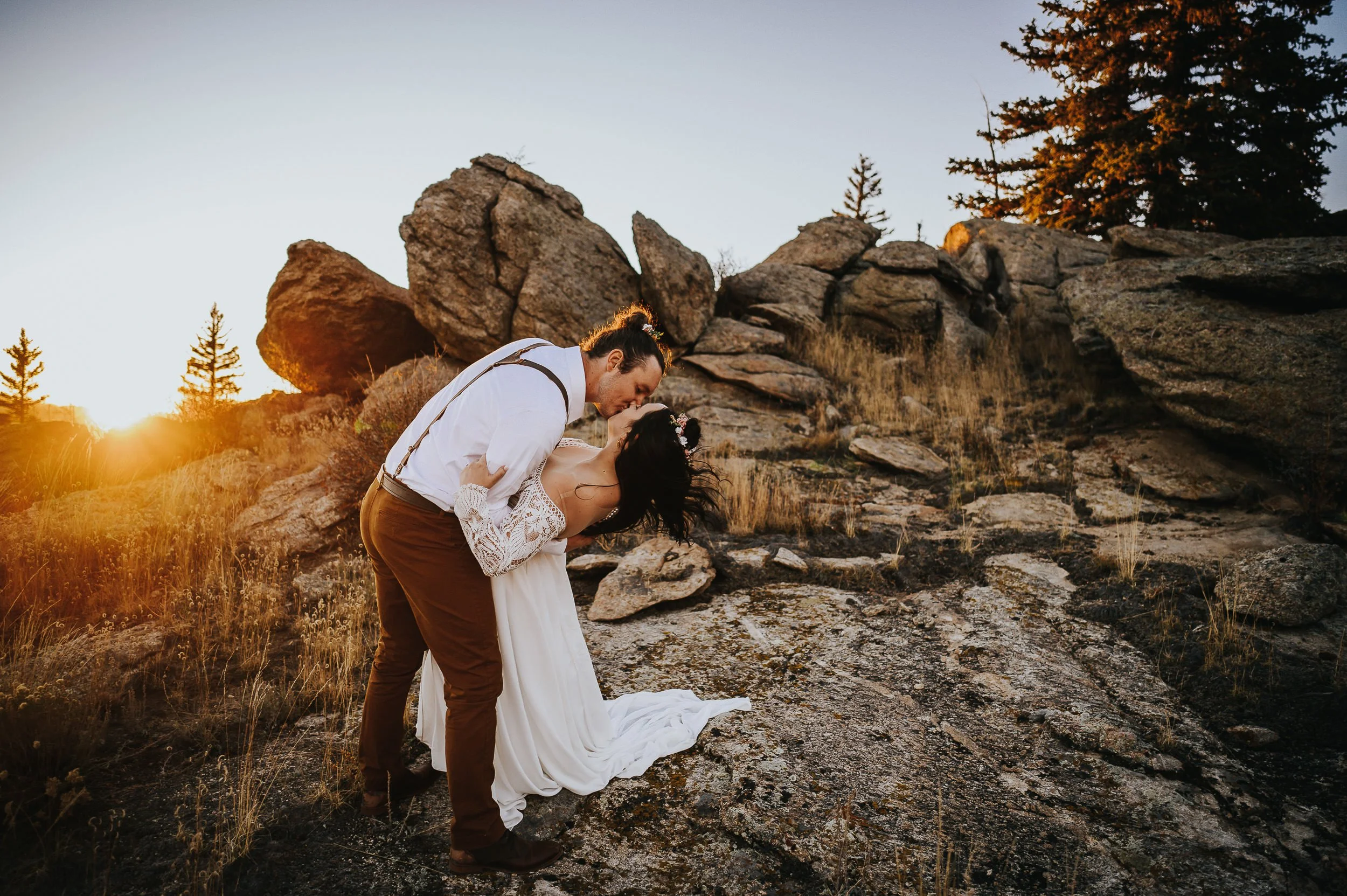 A couple kisses on top of a boulder at golden hour, Colorado mountains and forest stretching behind them.