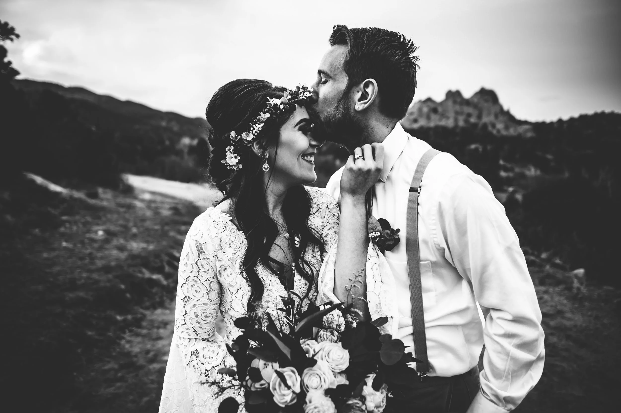 A bride and groom share a forehead-to-forehead kiss in a black and white portrait, she in a flower crown, he in suspenders.