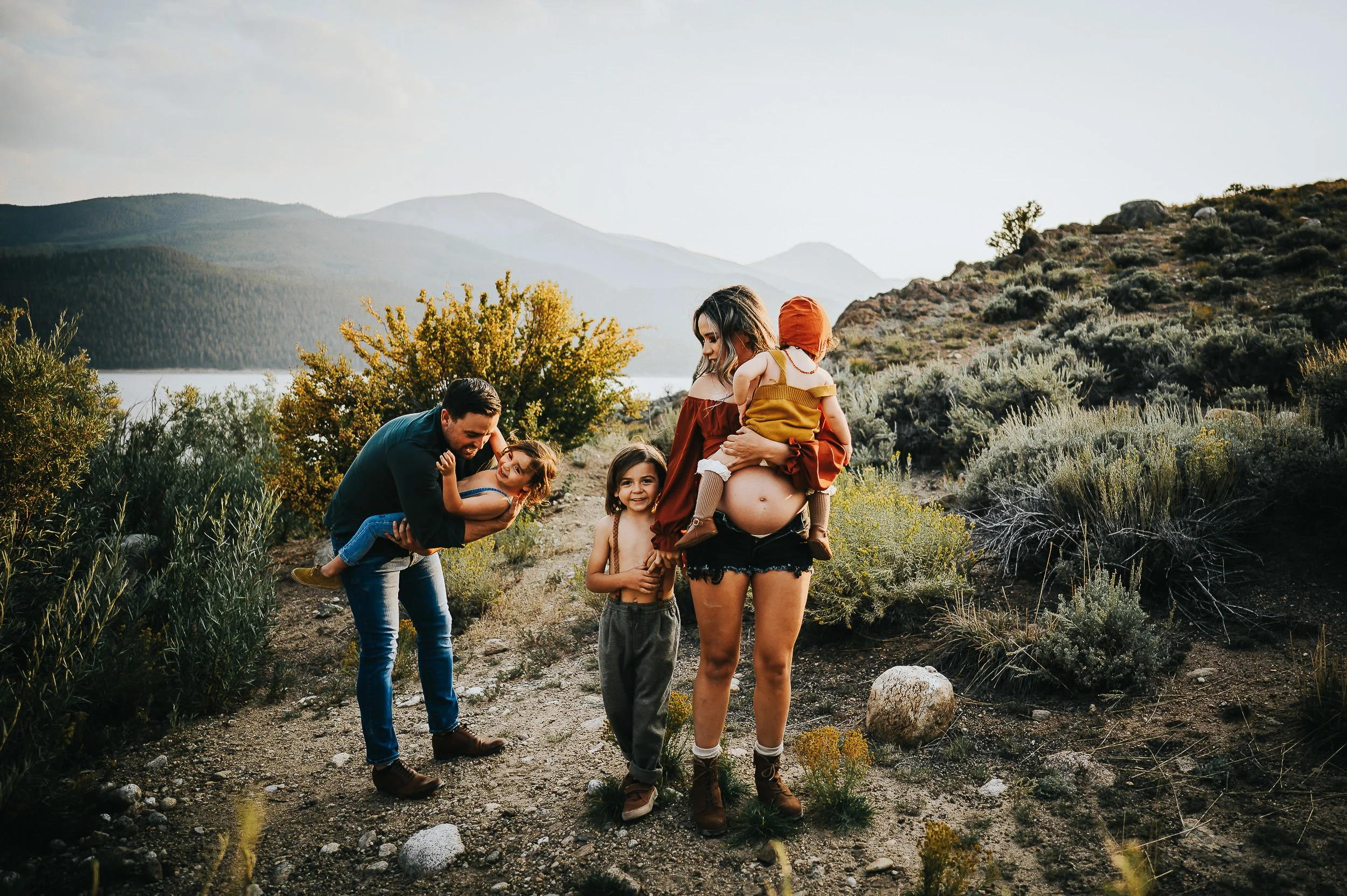 A family gathers in a Colorado wildflower field, dad leaning in to kiss a child while mom holds a toddler and shows her pregnant belly