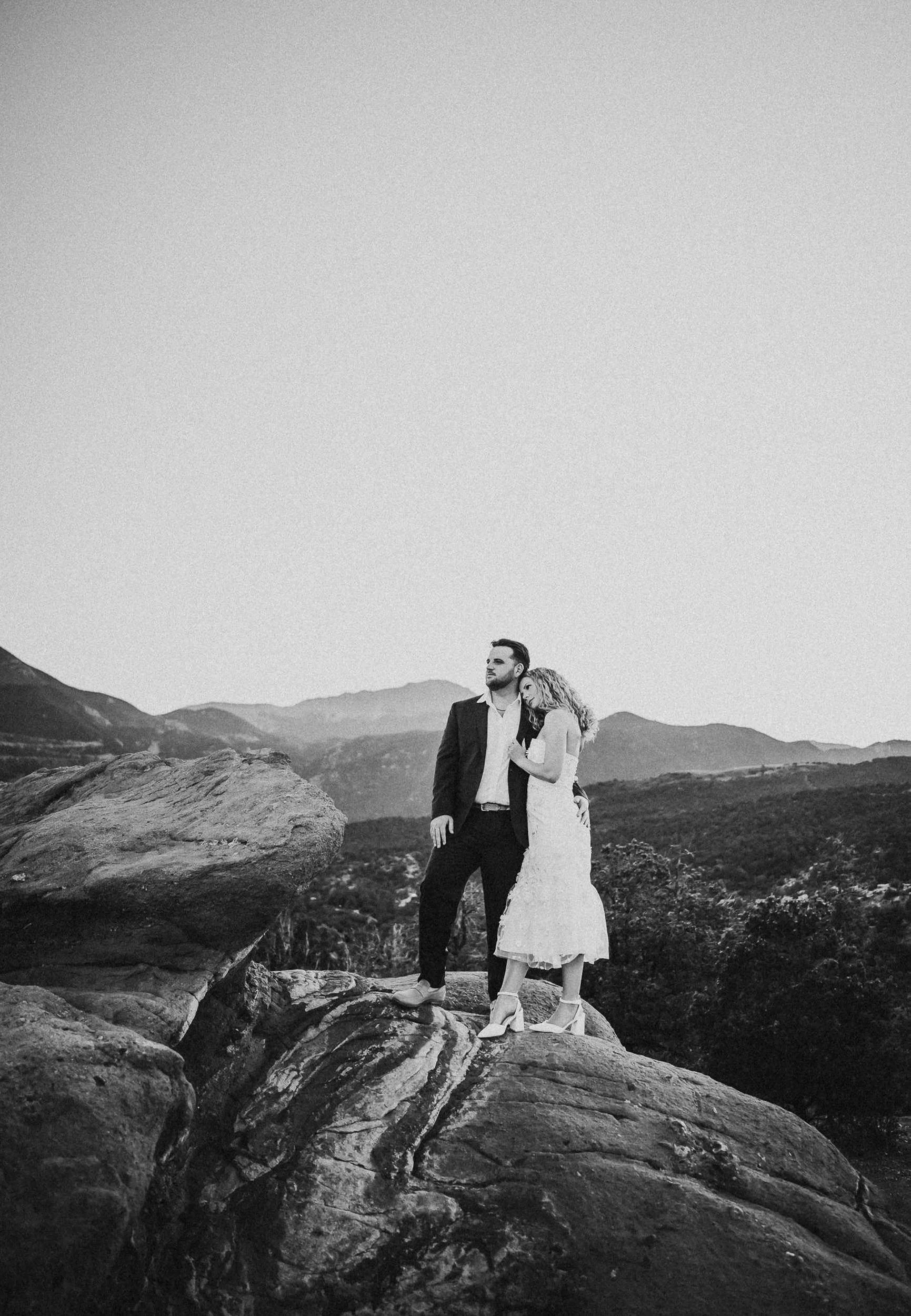 A bride and groom stand together on a rocky Colorado overlook in black and white, wide landscape shot.