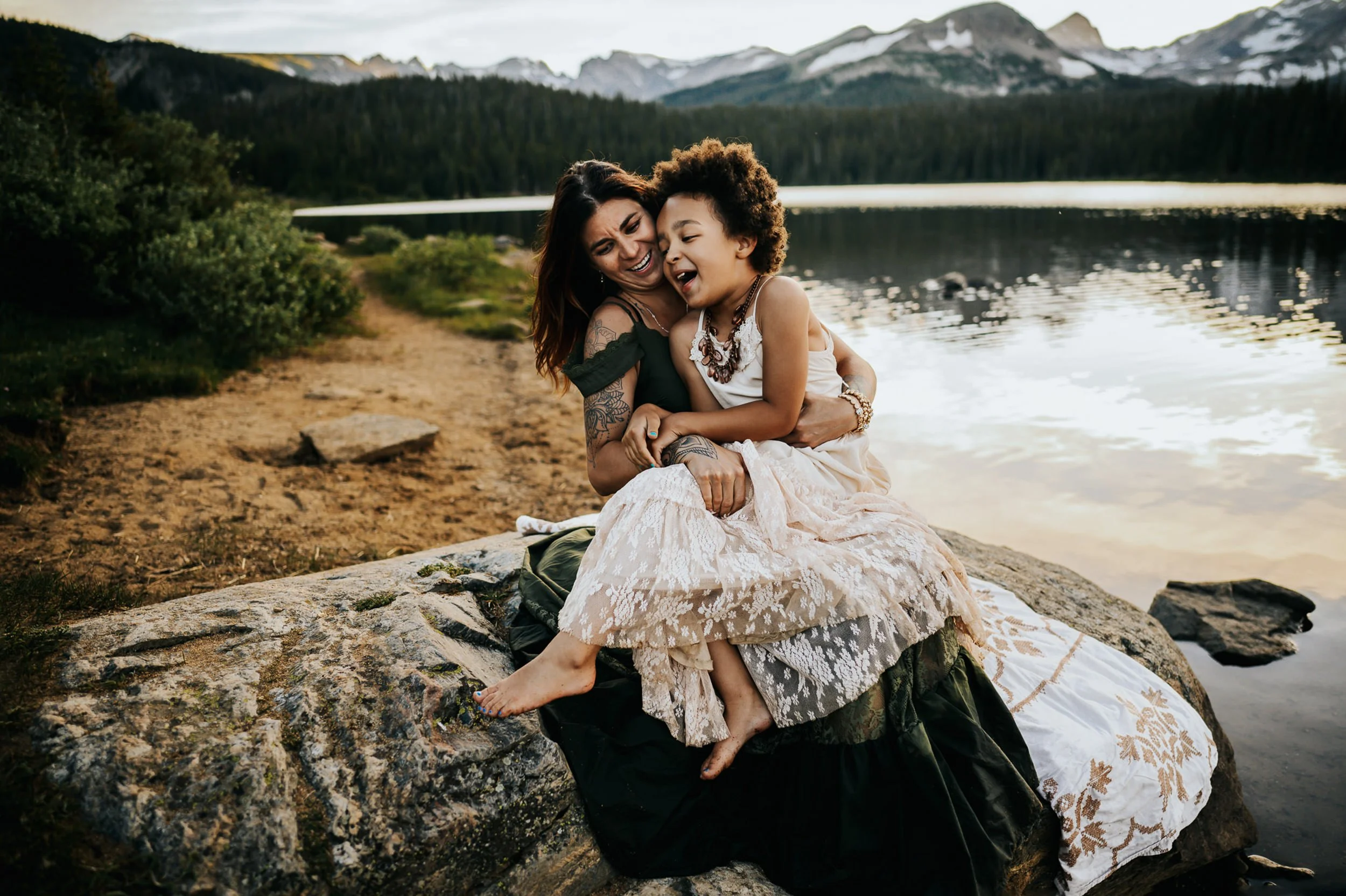 A girl is joyfully sitting with her mother near Brainard Lake, Colorado with the snow capped mountains towering behind them.