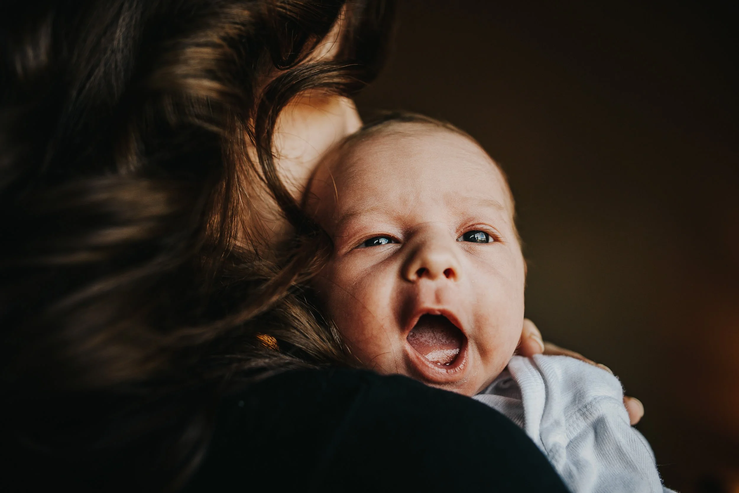 A baby yawns widely while looking over the shoulder of his mother in the window light.