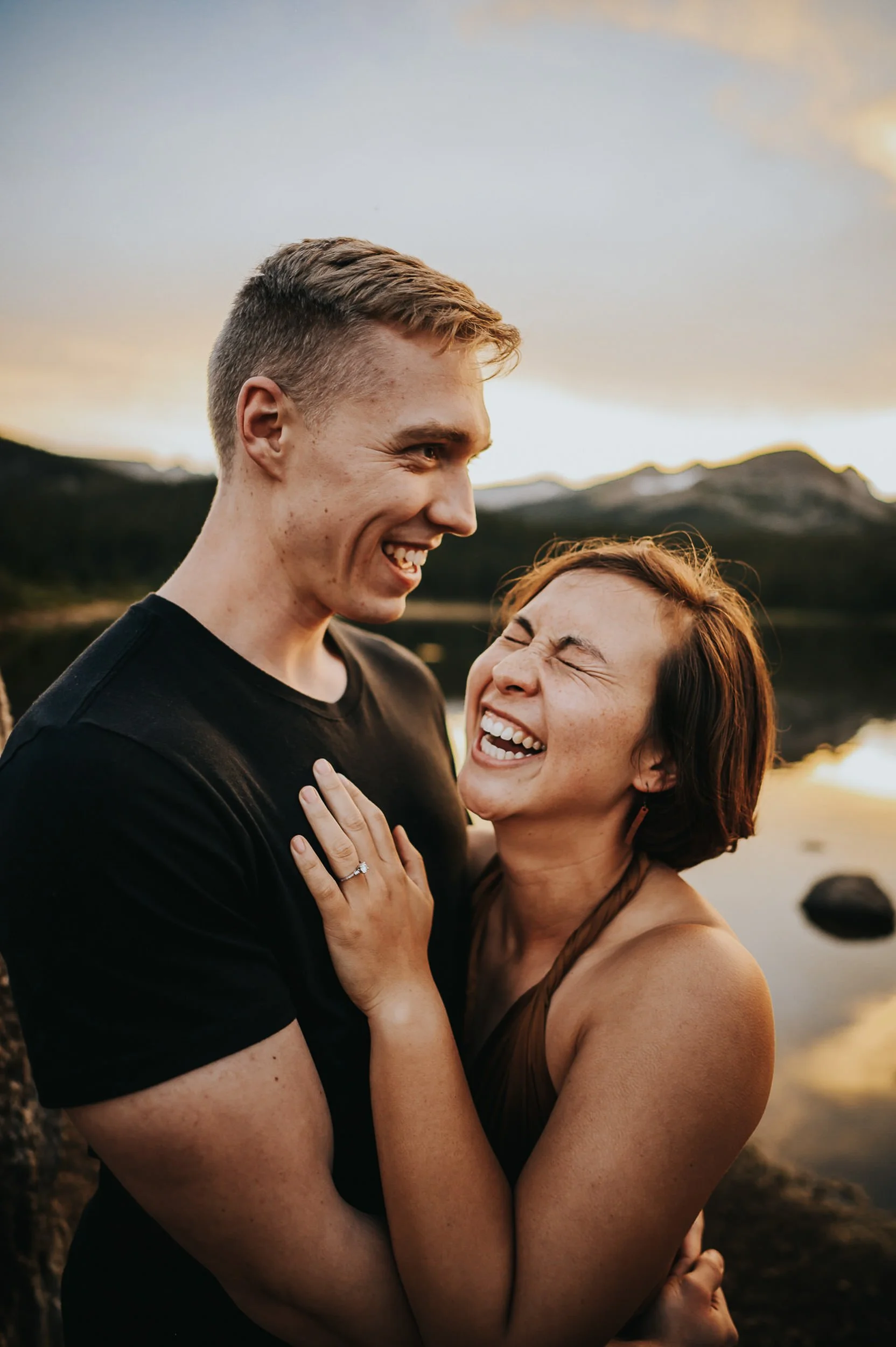 A couple laughs together beside a Colorado mountain lake, both facing each other with pure joy.