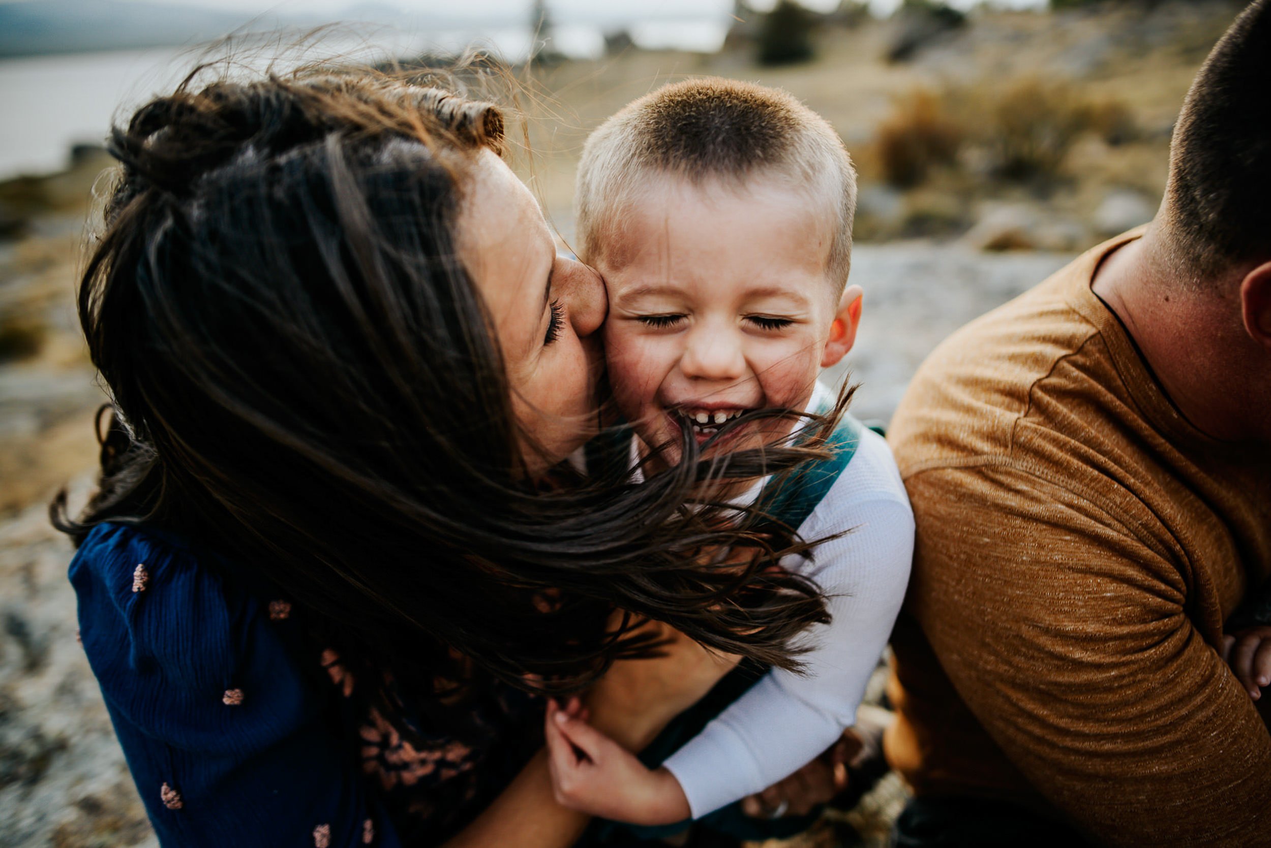 A parent leans in to kiss a laughing toddler's cheek while another parent looks on outdoors.
