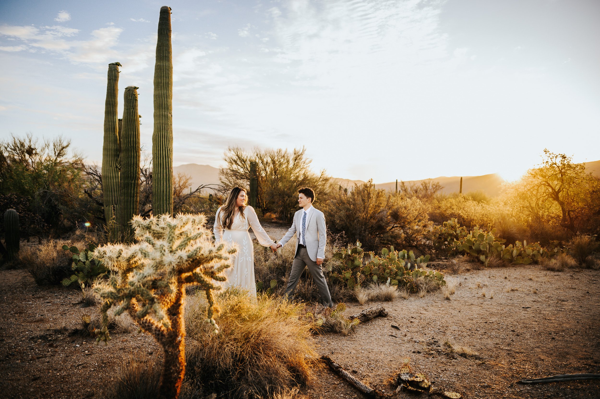 A couple stands together in a desert landscape at golden hour, tall saguaro cacti surrounding them.