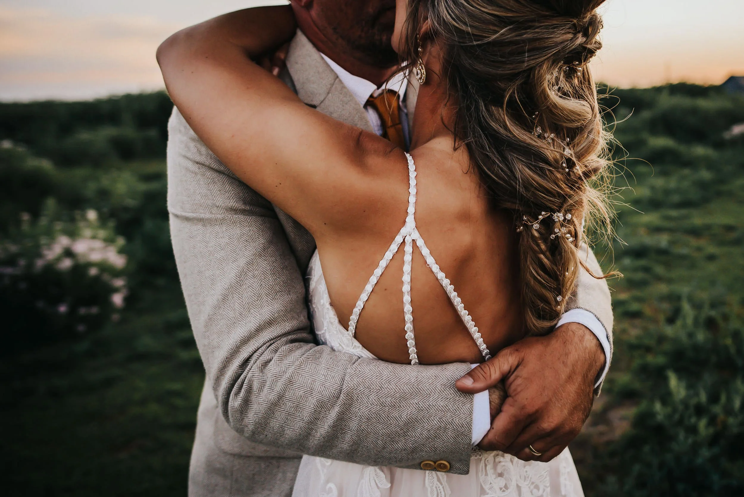 A groom wraps his arms around his bride from behind, her lace back detail visible, intimate closeup.
