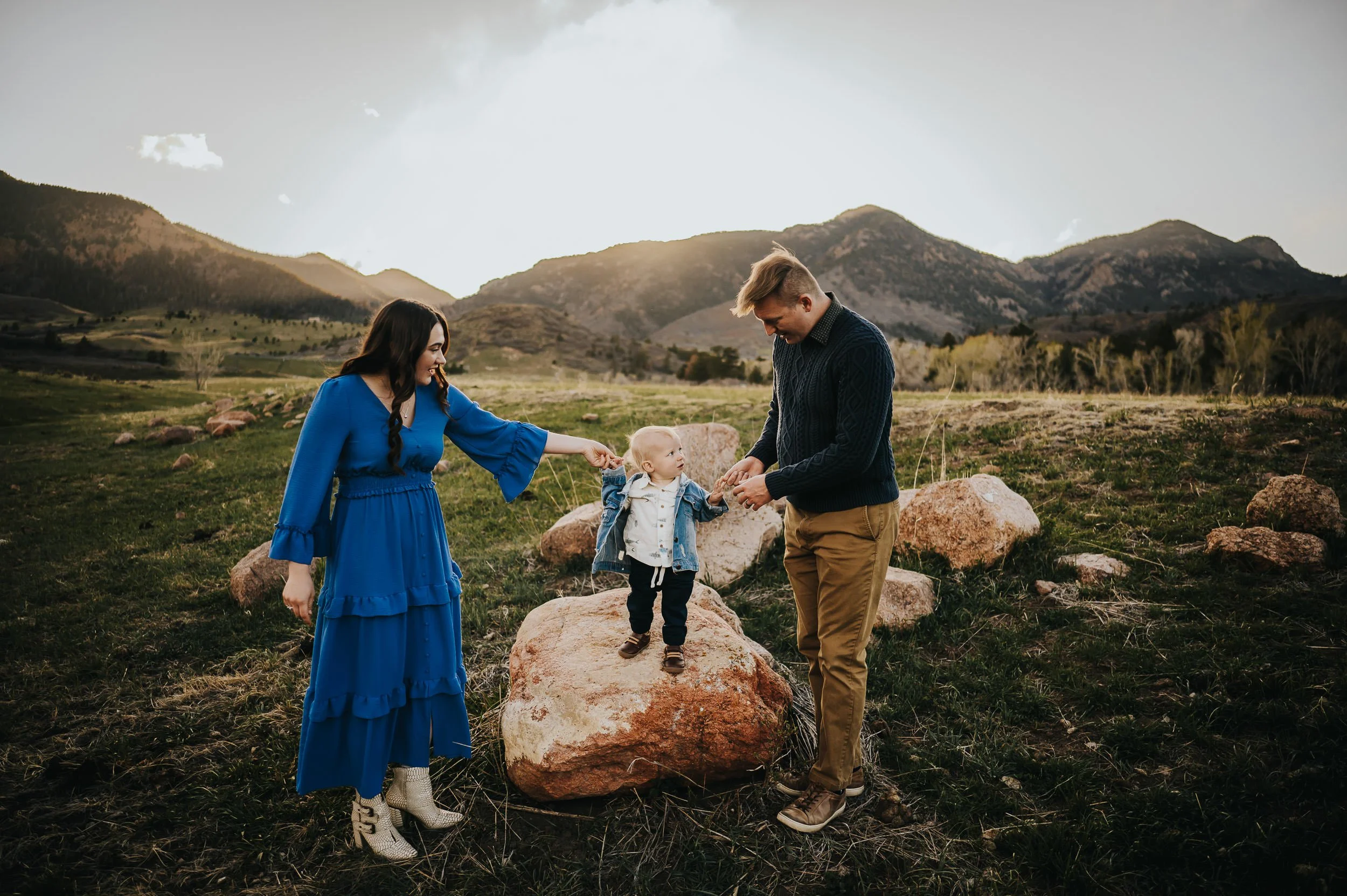 Family walking through a green mountain meadow in spring, mom carrying toddler, child running ahead, coordinated in soft earth tones.