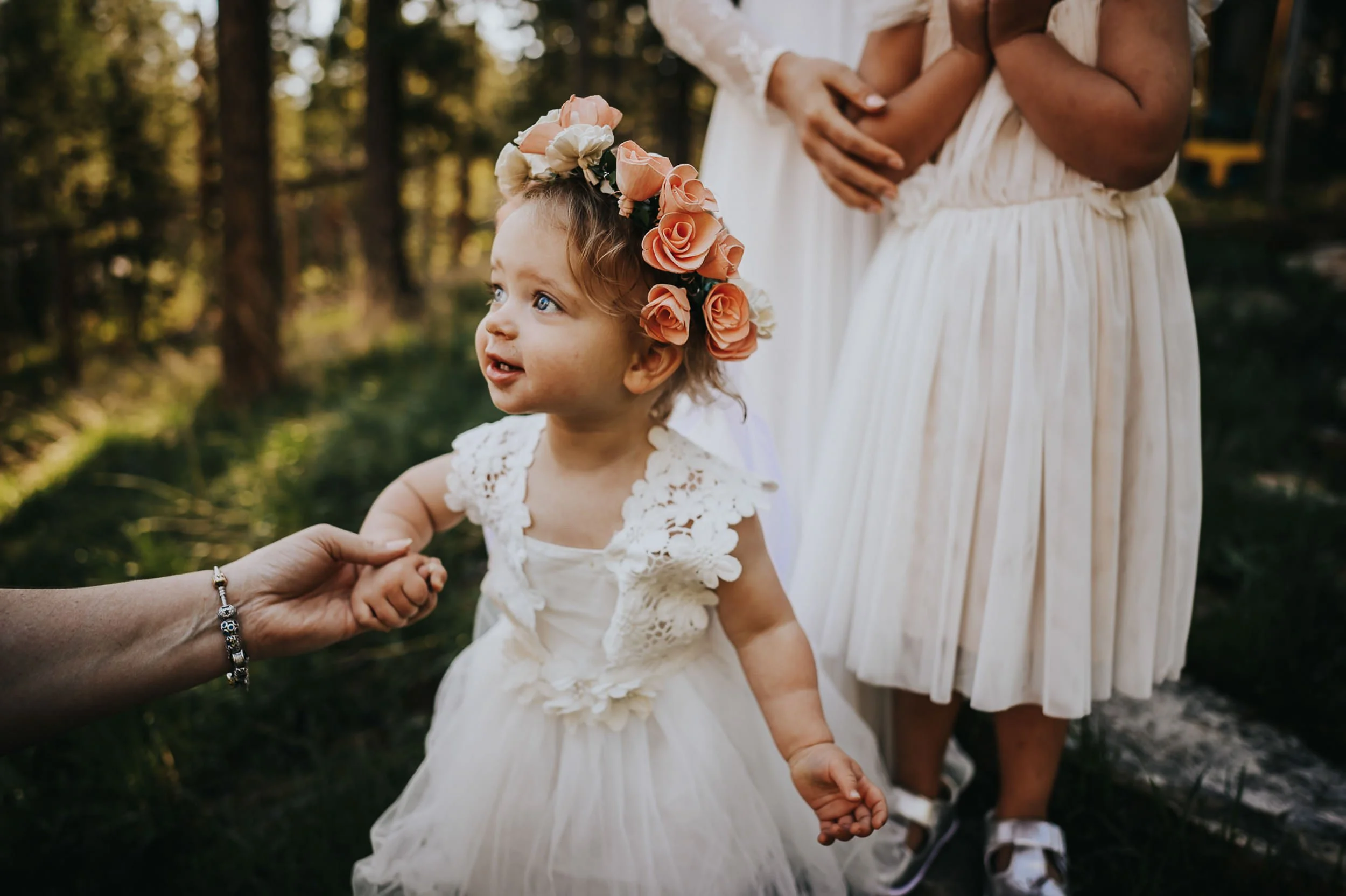 A little girl wearing a white dress and a rose flower crown holds the hand of an adult and smiles with the sun setting in the background.
