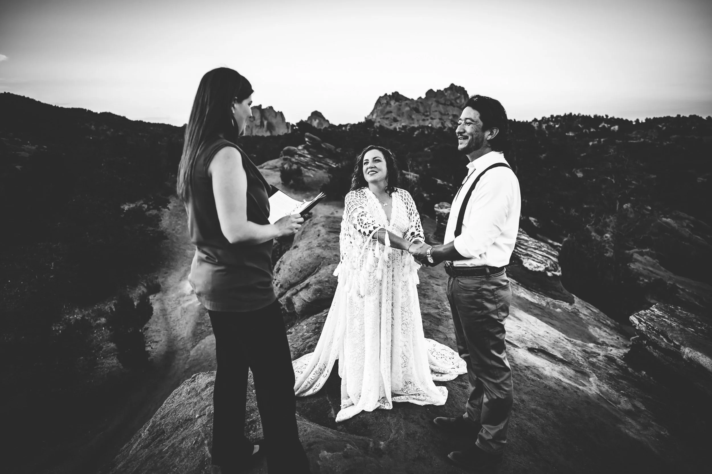 A couple exchanges vows in black and white at Garden of the Gods, officiant present, red rock formations behind them.