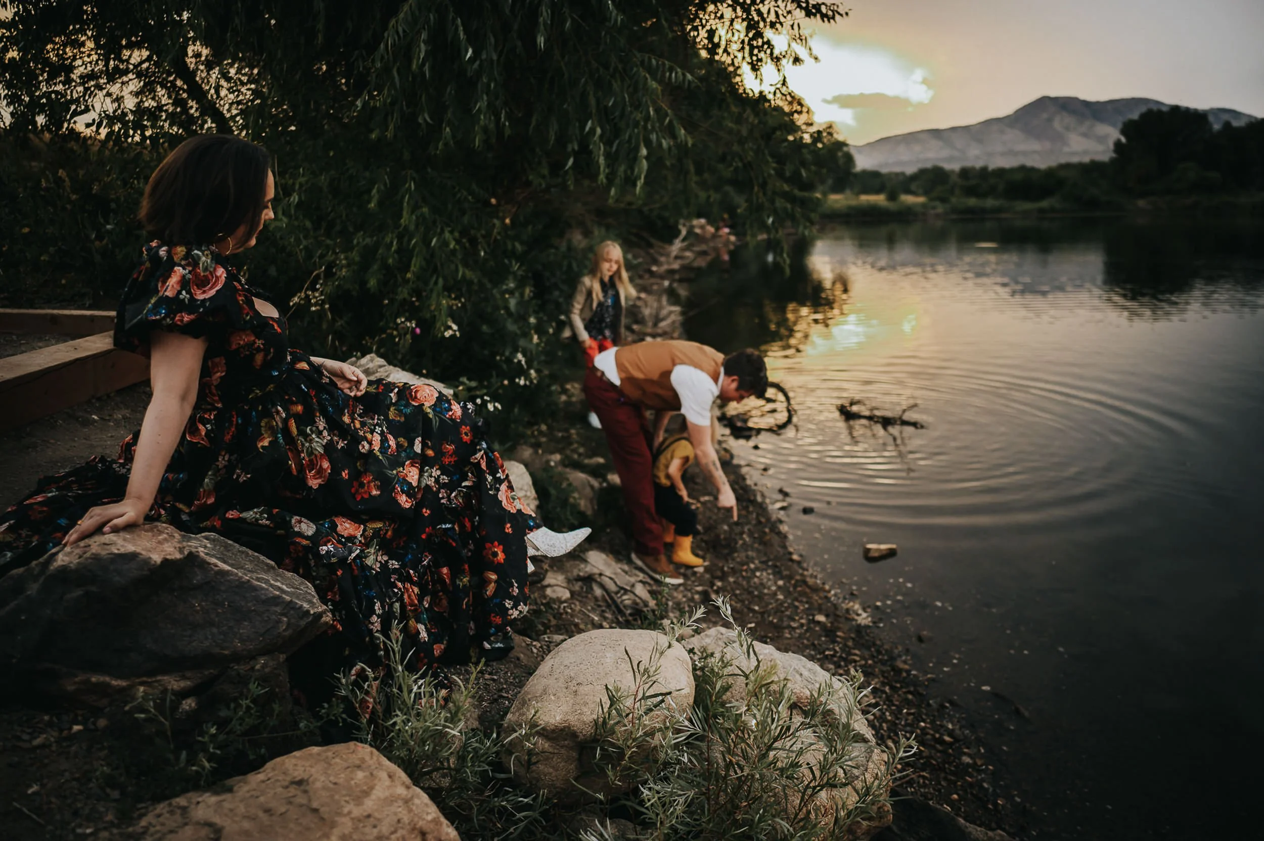 Mother wears a dark floral dress while she sits on a rock near a lake where her family explores.