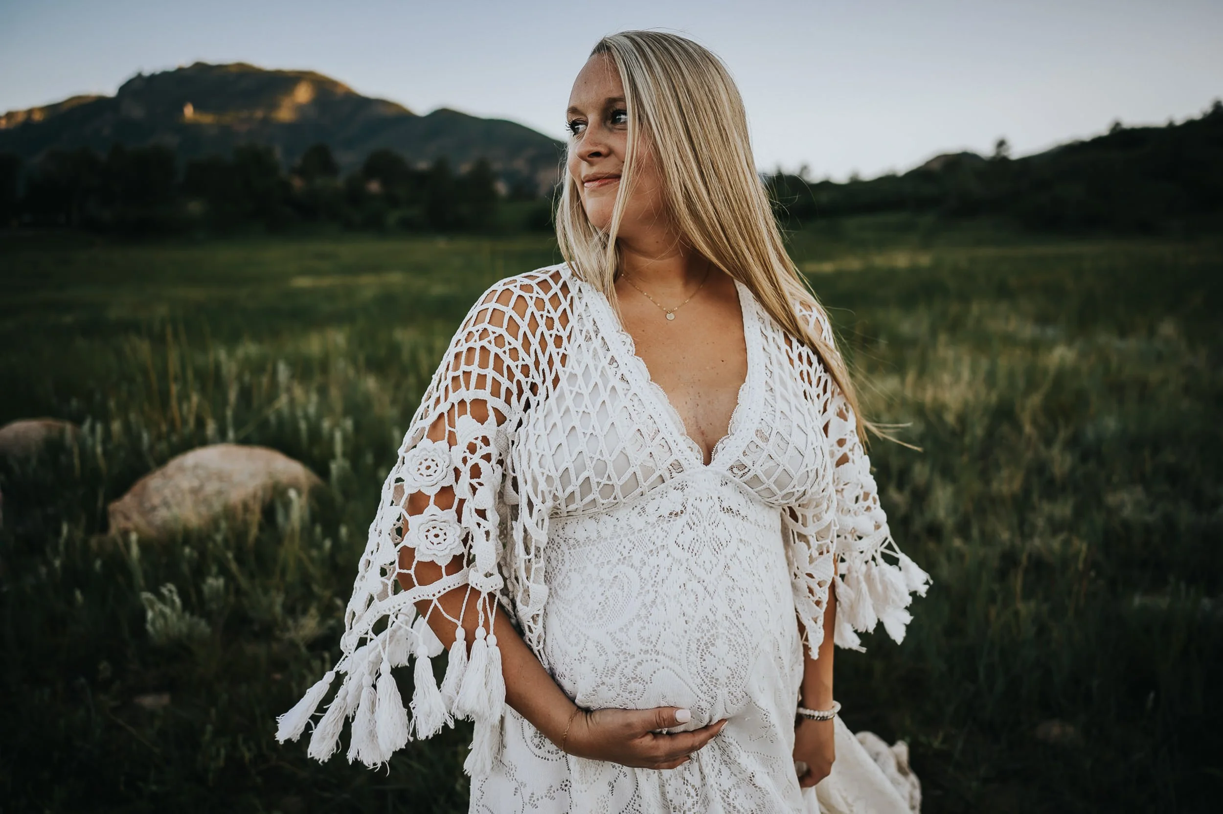 A pregnant woman twirls the bottom of her lace dress from Reclamation designs during her maternity session in a green field at Stratton Open Space in Colorado Springs.