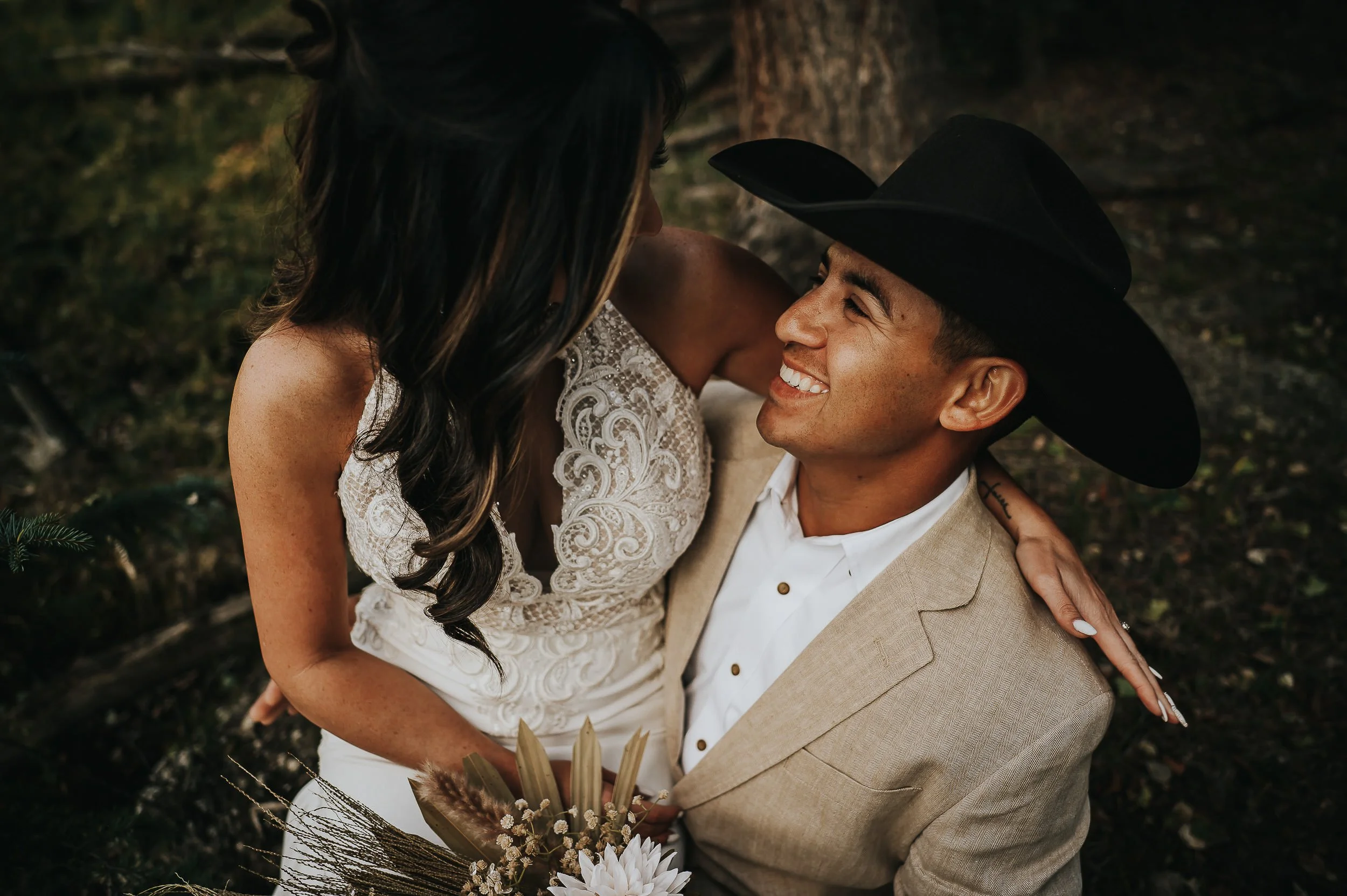 A man in a cowboy hat holds his partner close, both laughing warmly in an outdoor portrait.