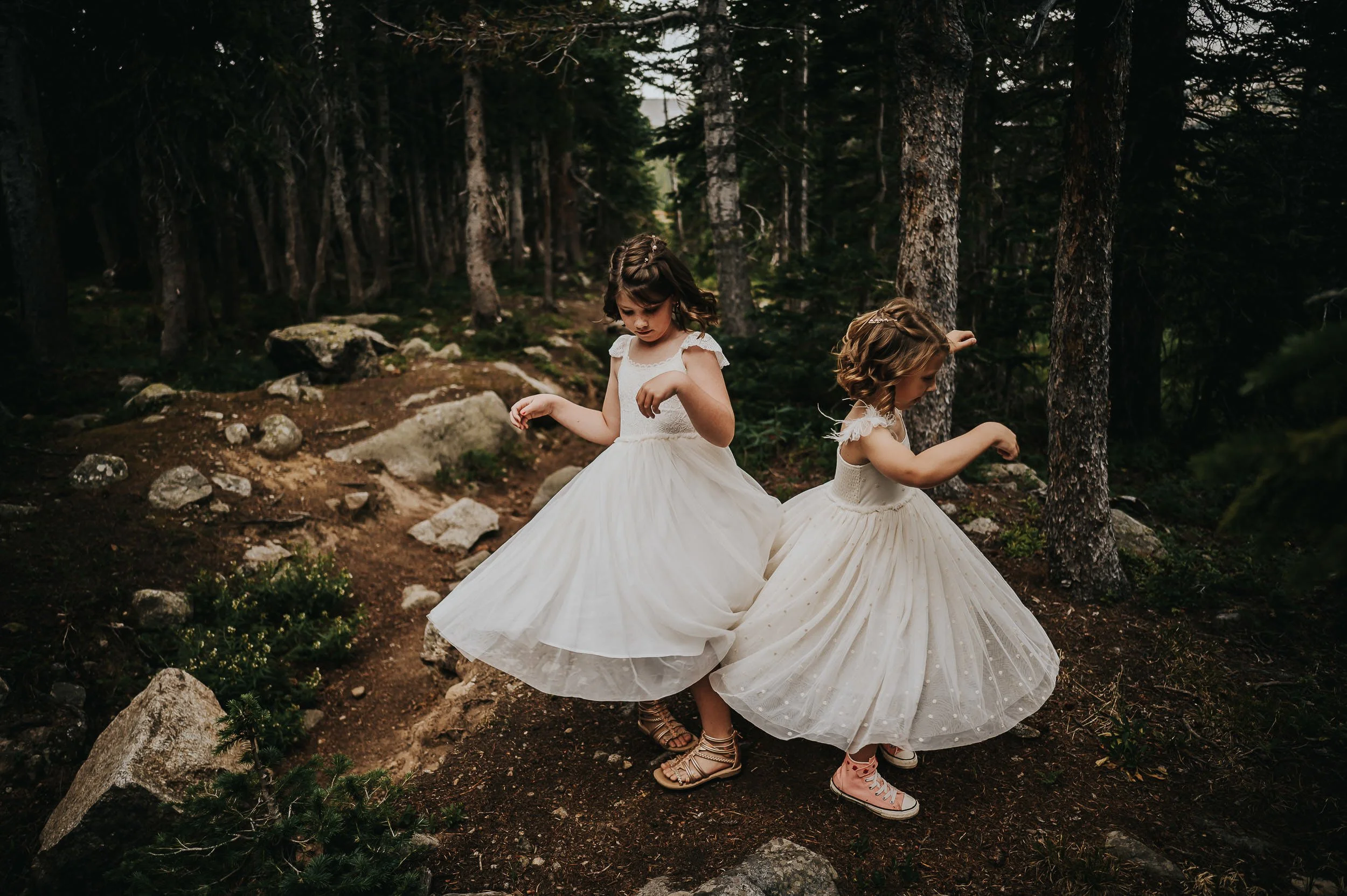 Two young girls in white dresses spinning and dancing together on a forest trail.